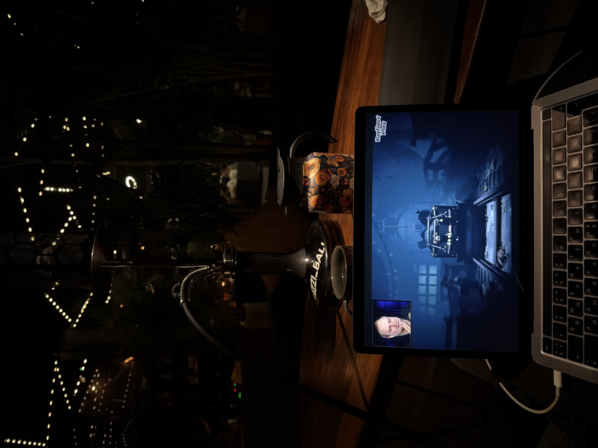 A dimly lit scene shows a laptop displaying a video of a man in the corner of the screen, apparently streaming a video game. The game has a dark and somewhat industrial-looking setting. A hookah sits beside the laptop on a wooden surface. In the blurred background, lights suggest an outdoor environment, potentially in a city. The keyboard of the laptop is visible, with keys displaying both English and Cyrillic characters. The hookah has a floral patterned bowl. Given the time on the screen and the context clues, it appears someone is enjoying the streaming video while using a hookah in a dimly lit area. No location can be confirmed other than what the image conveys. No data proves it's in North Kuta, Indonesia.