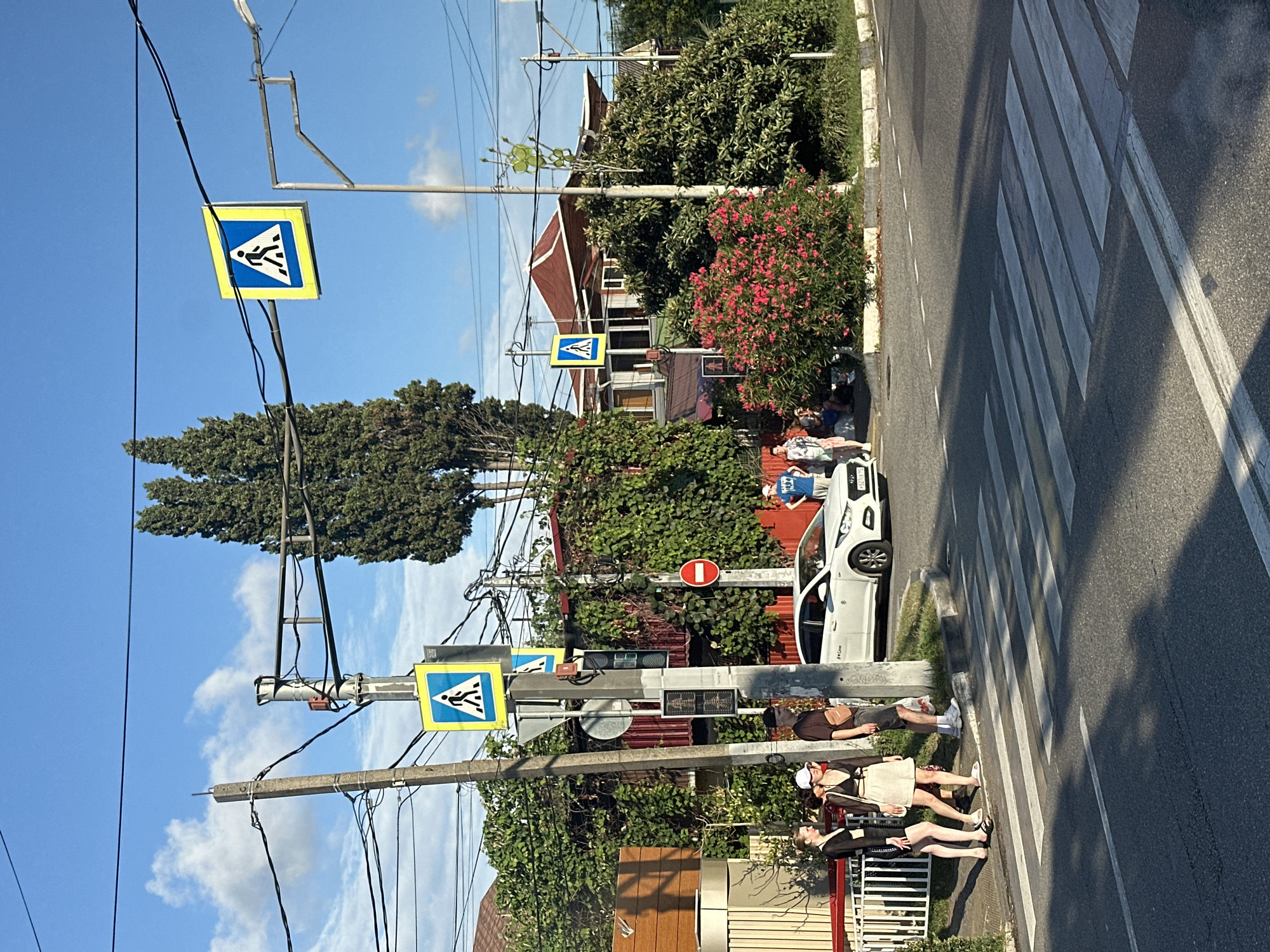 The image depicts a street scene in Sochi, Russia, at a pedestrian crossing. A white sedan is stopped at the crossing, indicating yielding to pedestrians. Several people are visible, with some standing near the crossing and others in the background. Two pedestrian crossing signs are prominently displayed on poles, along with traffic lights and other street signage. Lush greenery and trees surround the area, with residential buildings visible behind the foliage. The sky is blue with scattered clouds, suggesting a clear day. The overall impression is a typical urban environment during daylight hours.