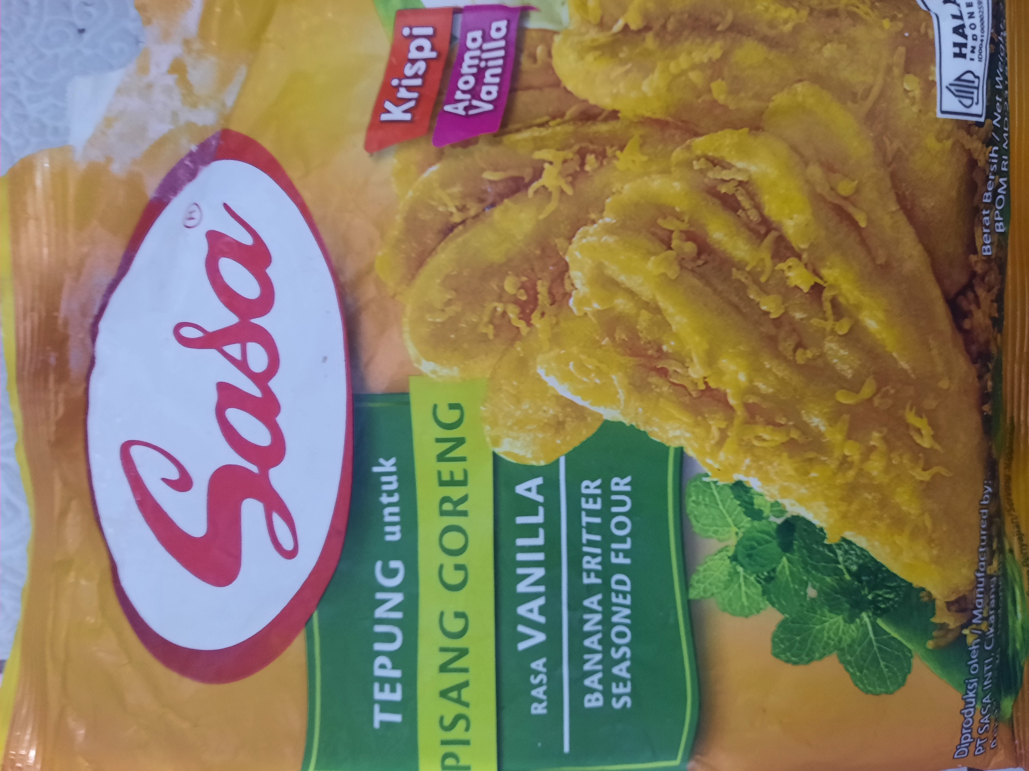 The image is a close-up, eye-level shot of a yellow package of "Sasa Tepung untuk Pisang Goreng Rasa Vanilla" or "Sasa Banana Fritter Seasoned Flour Vanilla Flavor." The packaging prominently features the red "Sasa" logo on a white oval with a red border. Below the logo, text in white outlines the product name in Indonesian, followed by "RASA VANILLA" and "BANANA FRITTER SEASONED FLOUR" in English. To the right, two small flags indicate "Krispi" and "Aroma Vanilla." The background shows a close-up of several fried bananas coated in batter, with some mint leaves on the left side. At the bottom of the package, there is text indicating "Diproduksi oleh / Manufactured by: PT SASA INTI, Cikarang" and "HALAL INDONESIA."
The image does not depict or provide any information related to the city of Lhokseumawe, Indonesia.