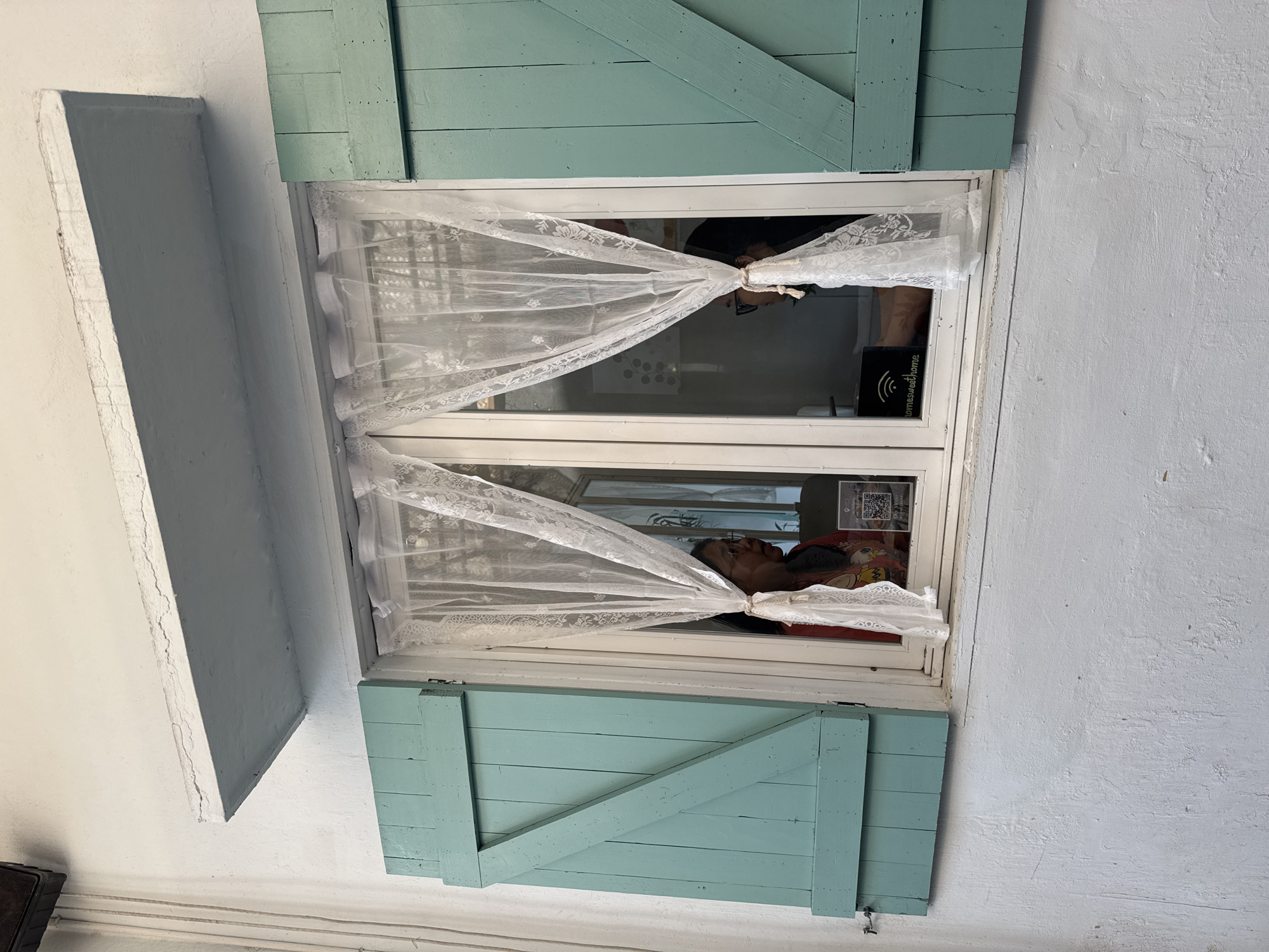 A window with light blue shutters is open and partially covered by sheer white curtains. Inside, a person with dark hair and skin is visible in the lower pane, lying down with a red and orange object nearby. In the upper pane, another person is visible, along with a small black device labeled "homesweethome" with a Wi-Fi symbol. The window is set into a white wall, with a projecting section of wall to the left. The image may have been taken in Đà Nẵng, Vietnam.