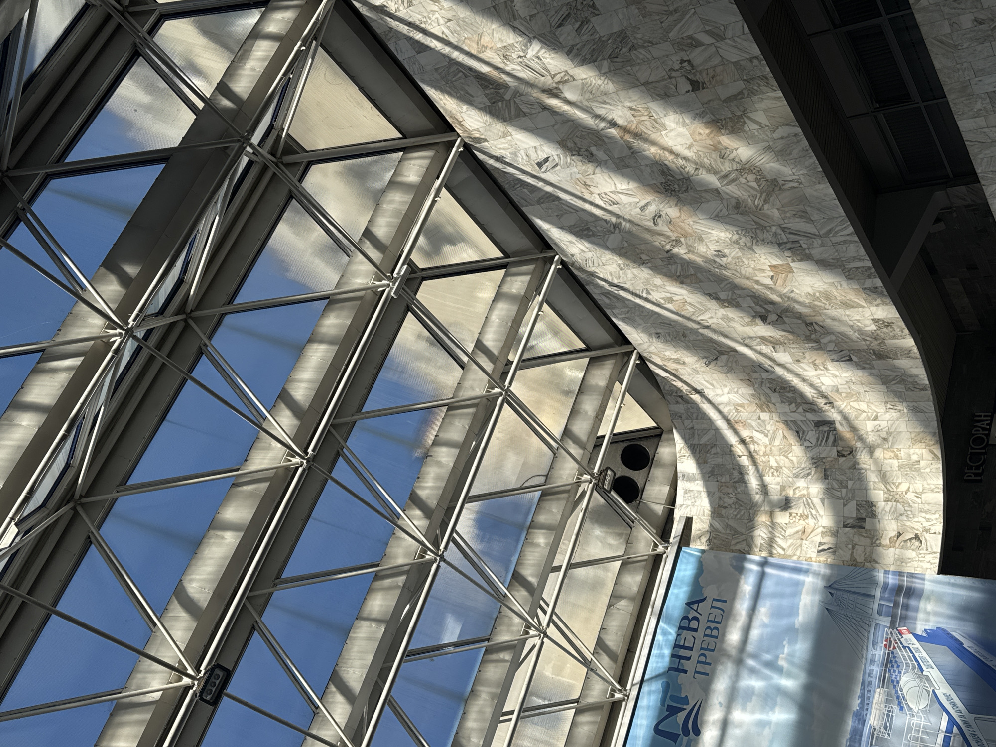 The image depicts an interior view of a building in Saint Petersburg, Russia, likely a shopping center or similar public space, characterized by modern architectural features.
The image is oriented in a way that the viewer is looking upwards towards the ceiling. The ceiling is composed of a grid-like metal framework holding large, transparent or translucent panels, allowing natural light to illuminate the space. The panels reflect a blue sky, suggesting it is daytime.
To the right of the grid structure, there is a curved section of the ceiling or wall covered in a light-colored, patterned tile or marble. Shadows cast by the grid structure form distinct, curved patterns on this surface.
At the bottom of the image, a portion of a promotional banner is visible. The banner appears to be for a tourism or travel company named "NEVA TRAVEL," featuring an image of a ship or boat. The text "РЕСТОРАН" is visible at the edge of the banner, suggesting it is in a restaurant.