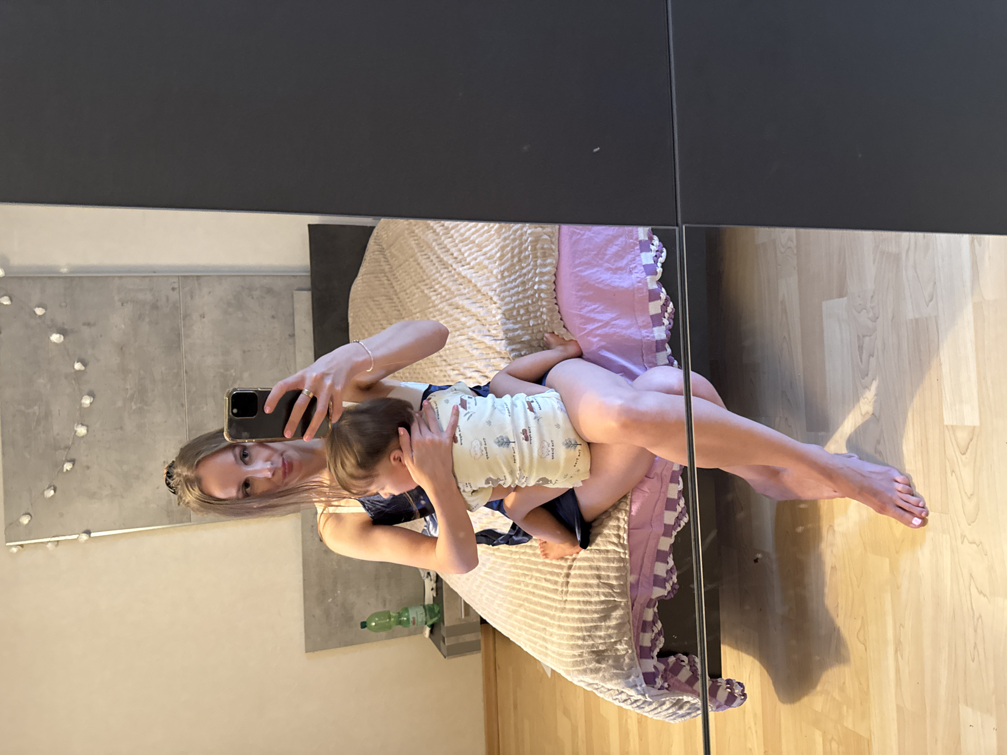 A young woman with long blonde hair is taking a selfie in a bedroom. She is sitting on a bed with her legs crossed, holding a baby on her lap. The baby is wearing a light-colored onesie with a sheep pattern. The woman is wearing a dark blue slip. The bed has a textured beige comforter and a pink sheet. A nightstand with a green bottle is visible to the left of the bed. The room has light-colored walls and wooden flooring. A large dark gray wardrobe is to the right of the scene. The image appears to have been taken in a residential setting in Überherrn, Germany.
