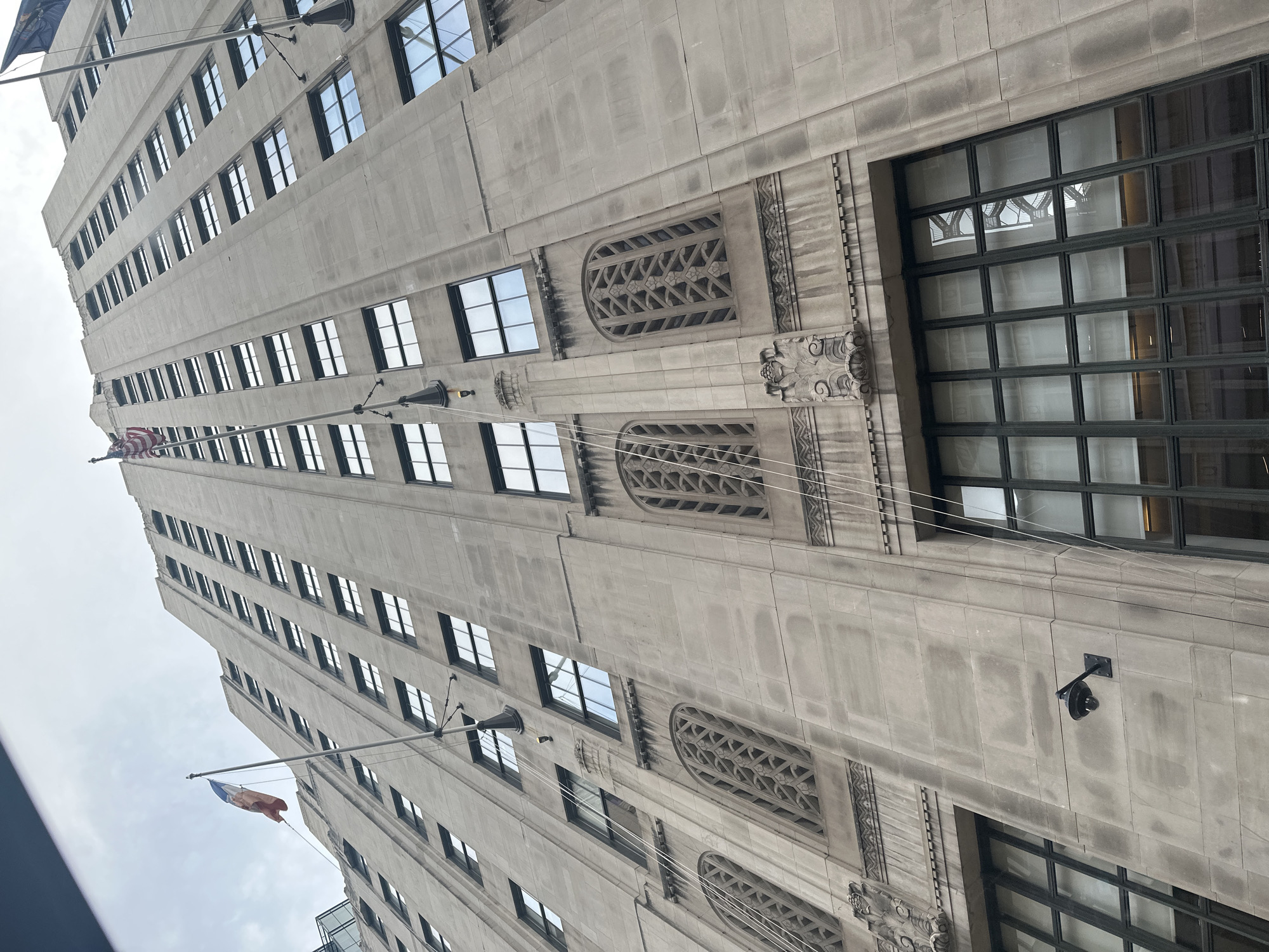 The image is a low-angle, worm's-eye view of the exterior of a tall, limestone-clad building, likely in New York City, United States of America. The building features a repetitive pattern of windows with dark frames, arranged in vertical rows. Some of the windows have blinds or curtains visible. Decorative elements, such as ornate carvings and arched window grilles, are present on the facade. Two flags are visible flying from poles attached to the building: one appears to be an American flag, and the other is a flag with blue, white, and red colors, possibly the French flag. There is also a security camera mounted on the building's facade. The sky above is overcast and cloudy.