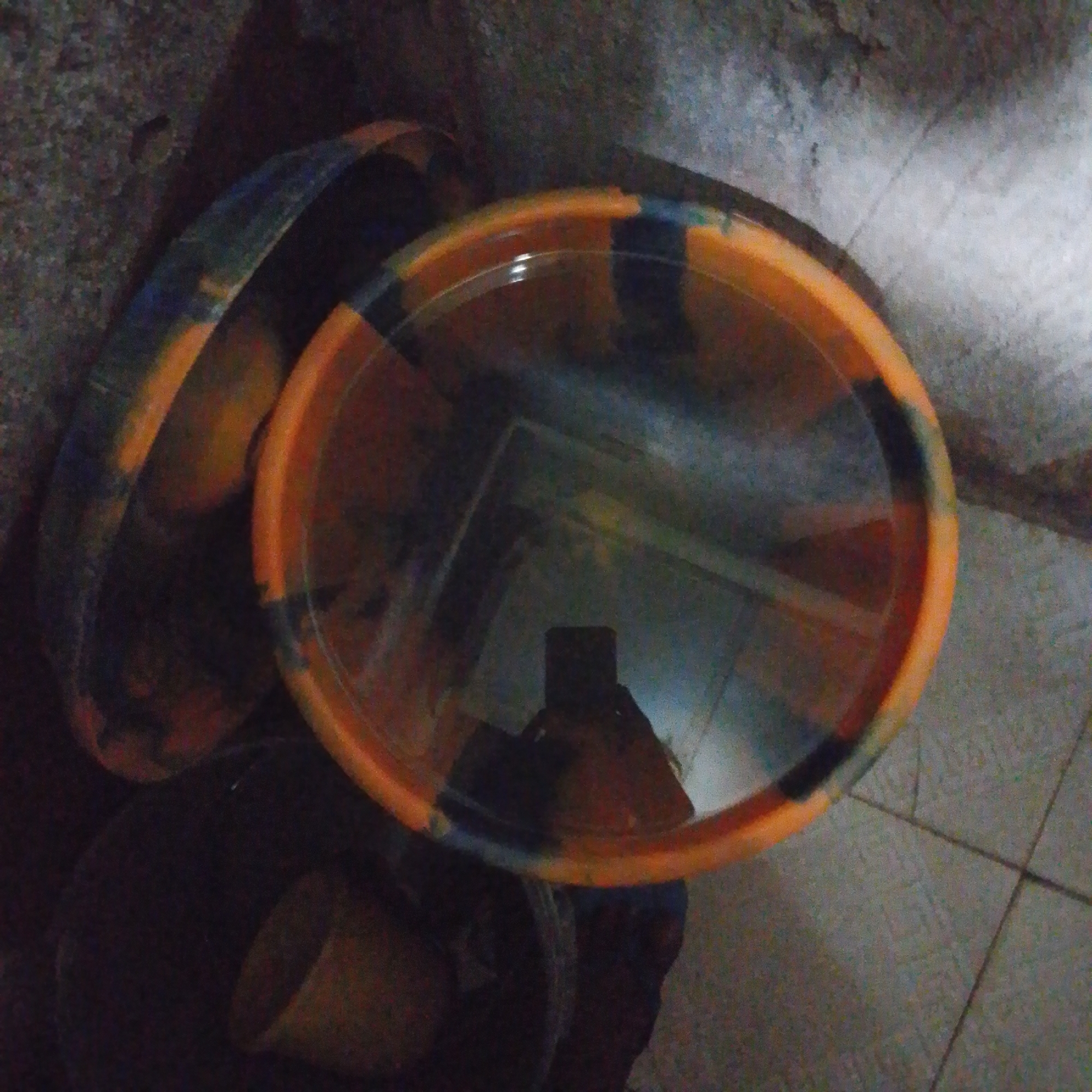 The image shows a close-up, overhead view of two stacked plastic basins with an orange, blue, and black marbled pattern. The top basin is larger and appears to be transparent with a reflection of a person holding a phone. The basins are resting on a textured, possibly concrete floor, with visible tile lines. The background is dark and appears to be a wall. The overall lighting is dim, suggesting an indoor or nighttime setting in Jalingo, Nigeria.