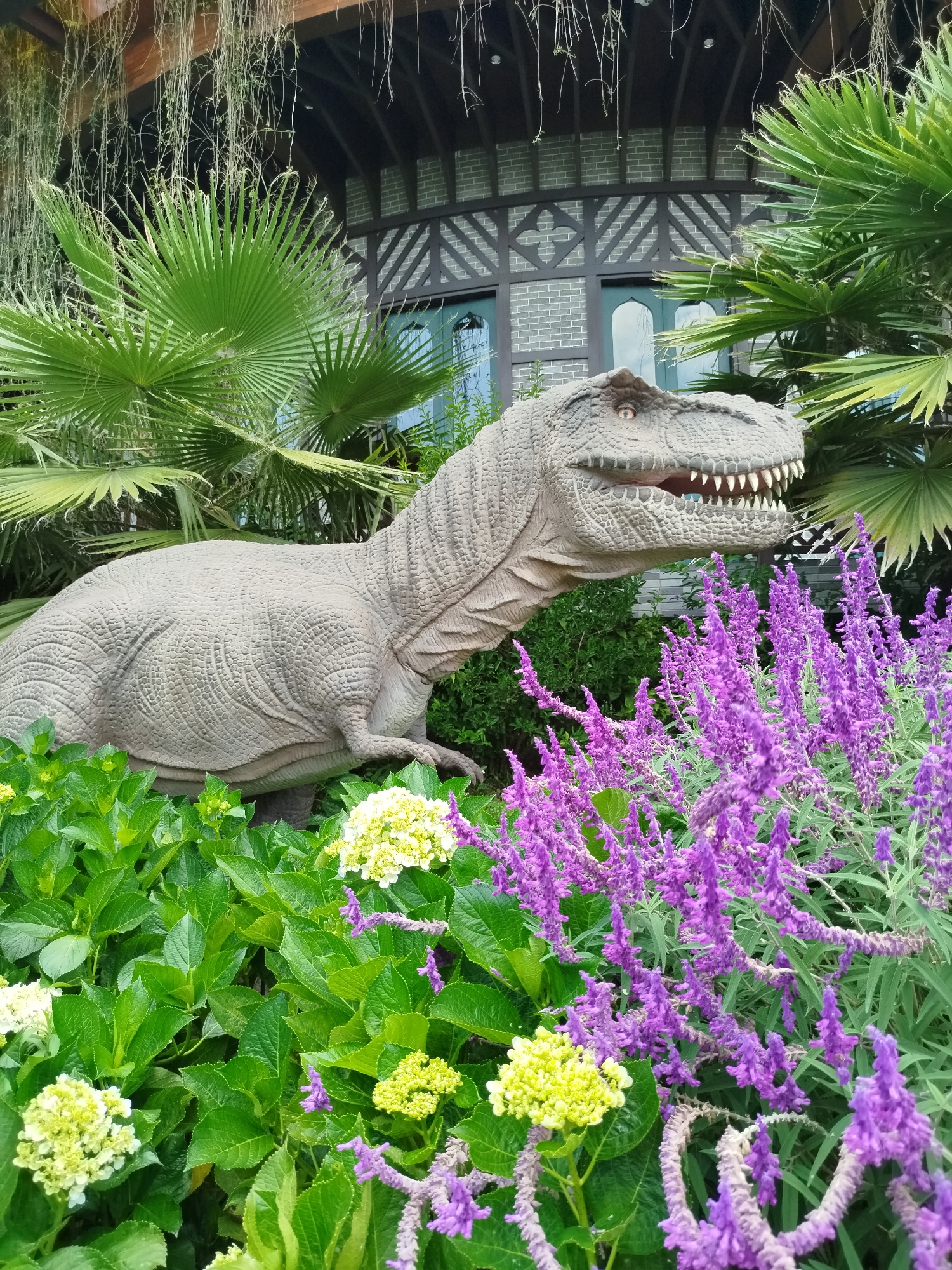 A full-length shot captures a T-rex dinosaur model in the gardens of the Fairy Garden in Cam Ly - Đà Lạt, Vietnam. The dinosaur, positioned slightly to the left of the frame, appears to be moving forward. The model is surrounded by plants and flowers, including palm trees, lavender, and hydrangeas. A building stands in the background, featuring a brick facade and ornamental dark wood trim.