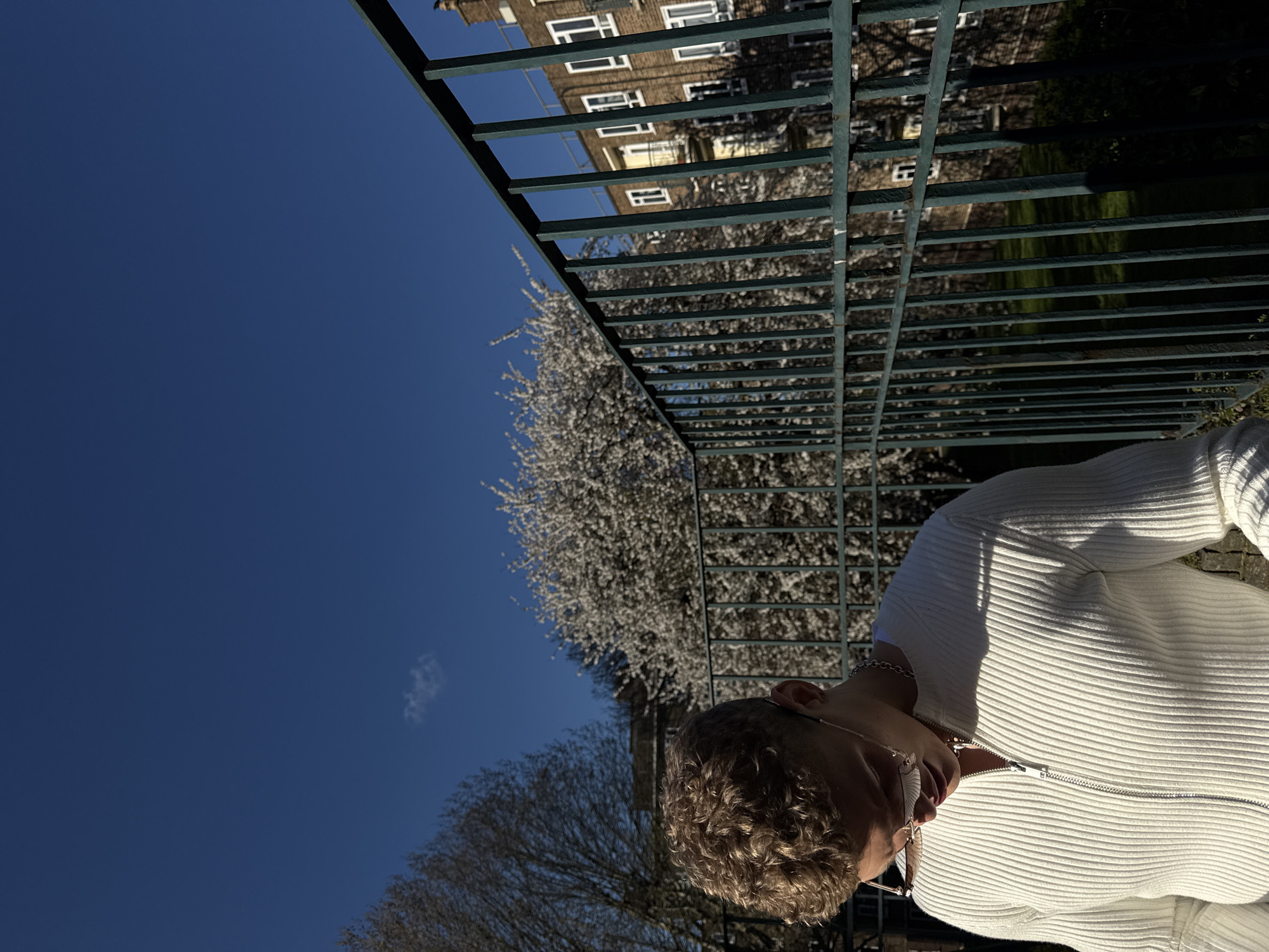 The image depicts a person with curly hair wearing a white zippered sweater and glasses, standing outdoors on a sunny day. The person appears to be standing near a green metal fence, with trees and a building visible in the background. The location is likely in London, United Kingdom, given the architectural style of the building and the type of trees. The sky is clear and blue.