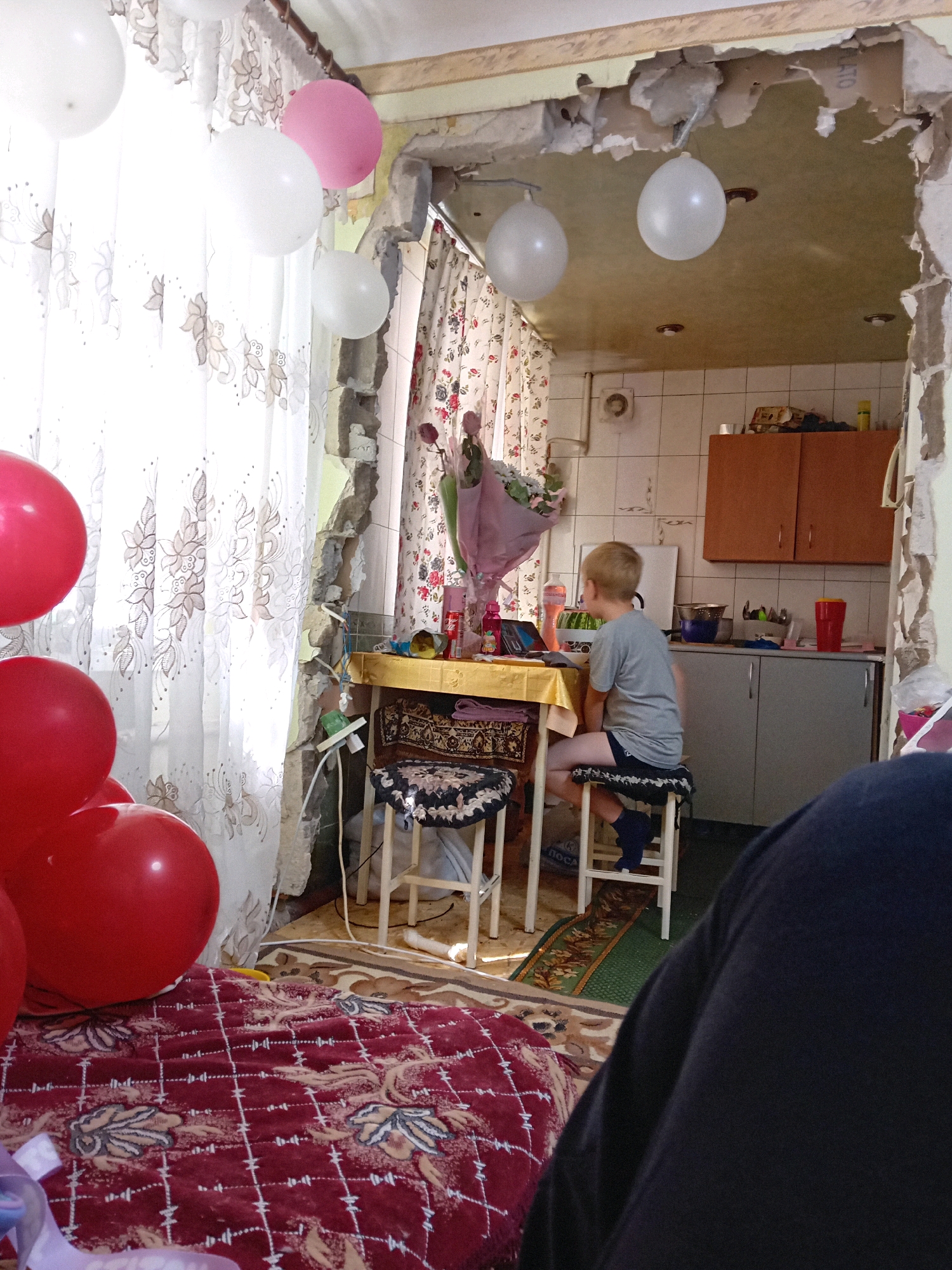 In a room undergoing renovation, a young boy with blond hair sits on a stool at a table, facing away from the camera. He is wearing a grey t-shirt and dark shorts. The room is decorated with several balloons, some white and pink, hanging from the ceiling. One wall appears to be demolished, revealing exposed concrete and wiring. The room contains kitchen cabinets, a sink, and a tiled backsplash. A patterned rug covers the floor. A person's leg is visible in the foreground, partially obscuring the view. The image is located in Chuhuiv, Ukraine.