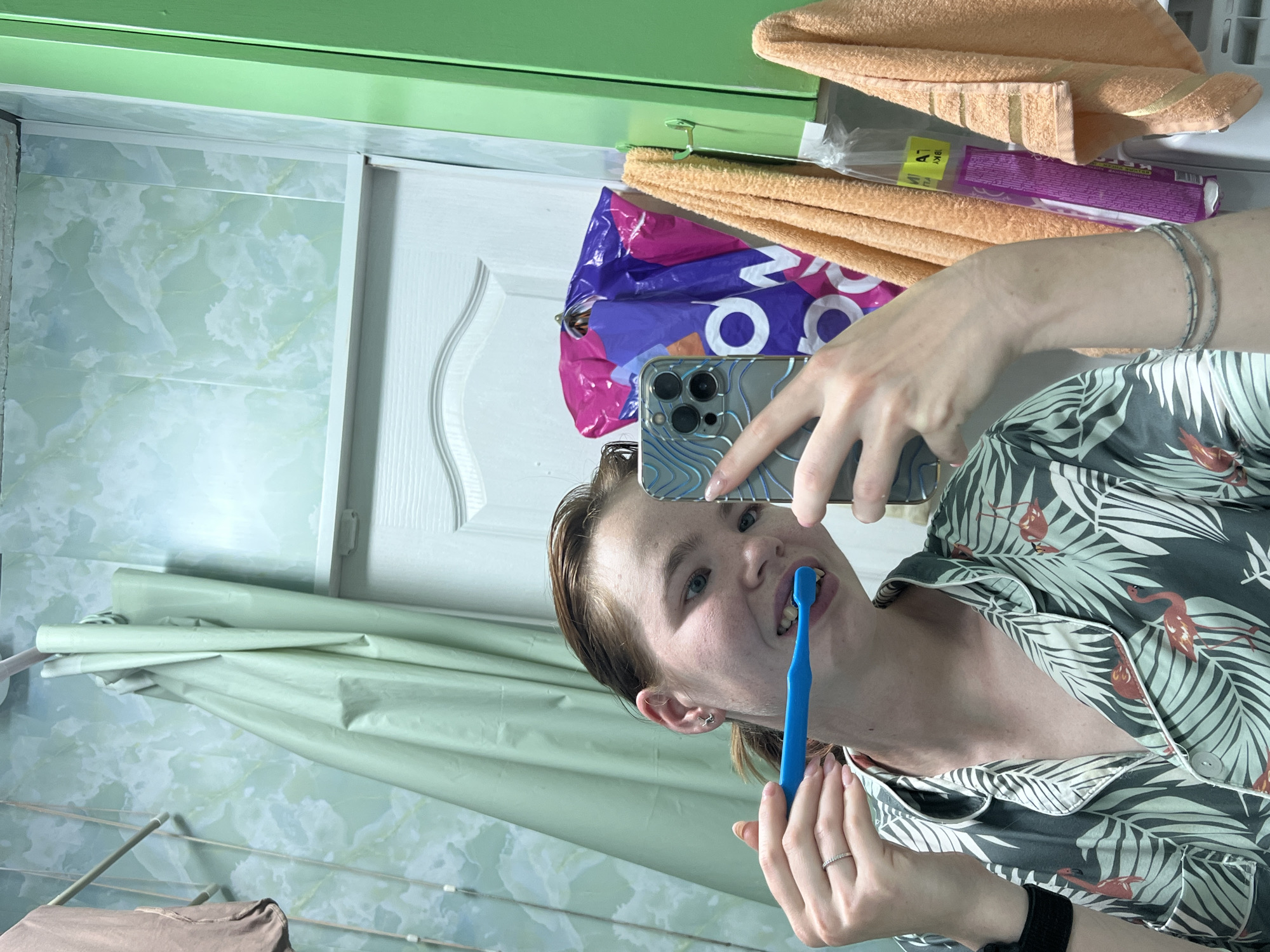 A young woman with light brown hair, pulled back in a ponytail, is taking a selfie in a bathroom. She is wearing a grey and white floral-patterned pajama top with flamingos on it. She is holding a blue toothbrush in her mouth and a smartphone in her right hand, which is taking the photo. Her left hand is holding the toothbrush. The bathroom walls are covered in a green and white marble-patterned wallpaper. A light green shower curtain is visible in the background, along with a white door. To the right of the woman, there are two orange towels hanging on hooks, and a purple shopping bag with white letters is also hanging on a hook. A thin silver bracelet is visible on her left wrist. The scene appears to be in a residential bathroom. The image was taken in Chaikovsky, Russia.