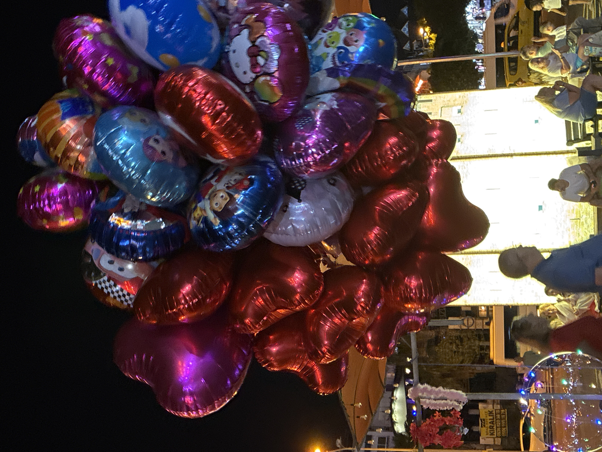 A cluster of colorful balloons floats in the night sky in Alanya, Turkey, near a street with people and vehicles. The balloons are varied in shape and design, with some appearing to be heart-shaped and others oval. Several balloons feature cartoon characters and patterns like checkerboards and stars.
Below the balloons, a street scene unfolds. People are seated on benches, some appearing to be locals and tourists. A yellow taxi is visible on the right side of the image, suggesting a busy urban environment. On the left, a building with lights and signage indicates a commercial area, possibly with shops or restaurants. A streetlight illuminates the scene, casting a warm glow on the surroundings. The overall atmosphere suggests a lively evening in Alanya.