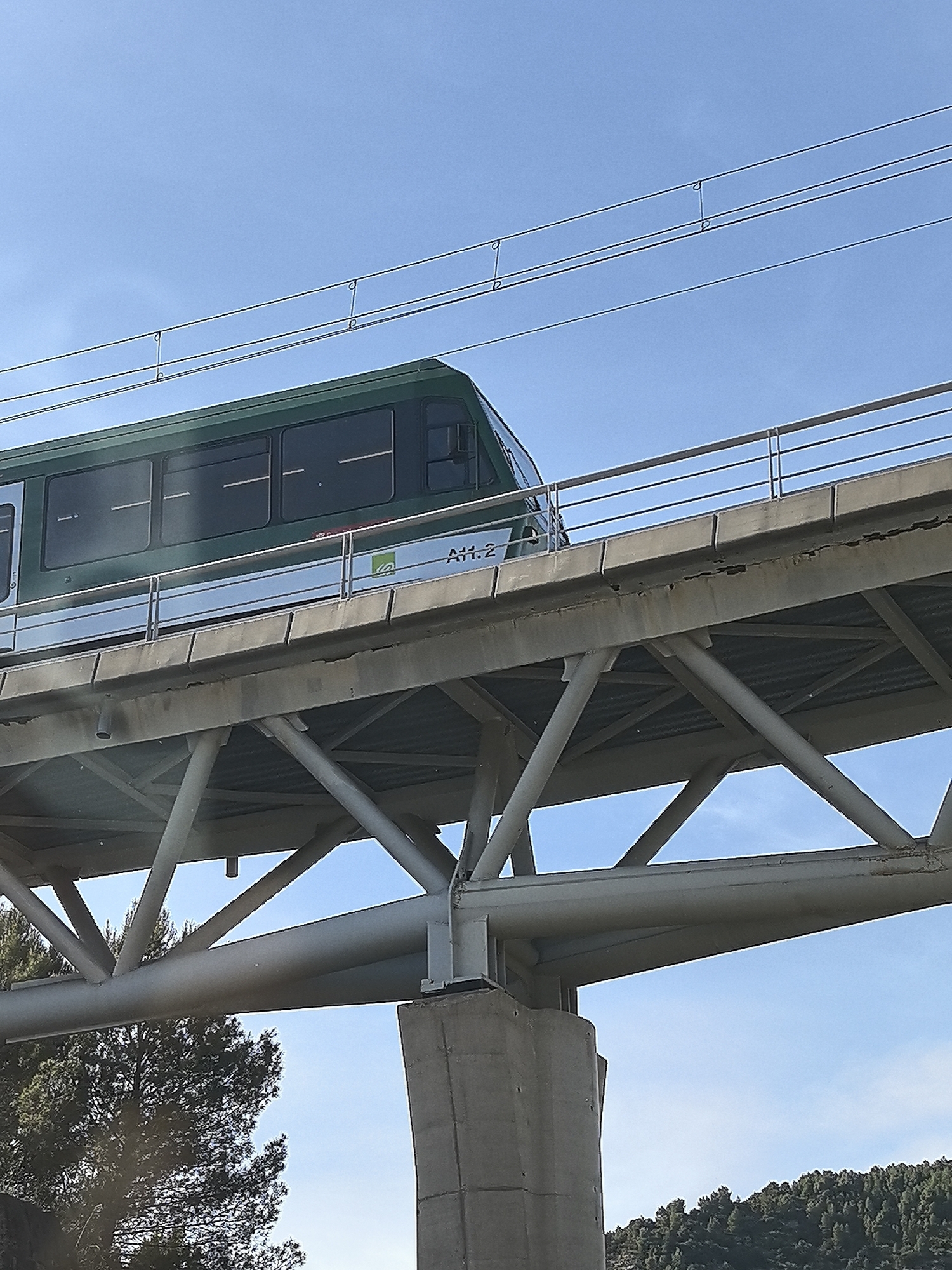 The image captures an elevated green and silver train, resembling a monorail or a rack railway car, positioned on a substantial concrete and steel bridge structure against a bright, clear blue sky. The train features multiple dark, rectangular windows along its side and has the alphanumeric identifier "A11.2" along with a small green logo visible near its front. Overhead, several parallel power lines run above the track.
The train is supported by an elevated concrete track bed, which includes a silver metal railing. Beneath this, a robust grey steel truss structure forms the bridge's main support, featuring numerous interconnected beams and diagonal braces. This steel framework rests on large, sturdy concrete pillars.
In the background, under the bridge and extending towards the horizon, are dense green trees covering what appear to be rolling hills or mountainous terrain. The bright sunlight indicates it is daytime, with no clouds visible in the sky. No people are present in the scene. Given the provided location context of Monistrol de Montserrat, Spain, this structure is likely part of the Cremallera de Montserrat (Montserrat Rack Railway).
