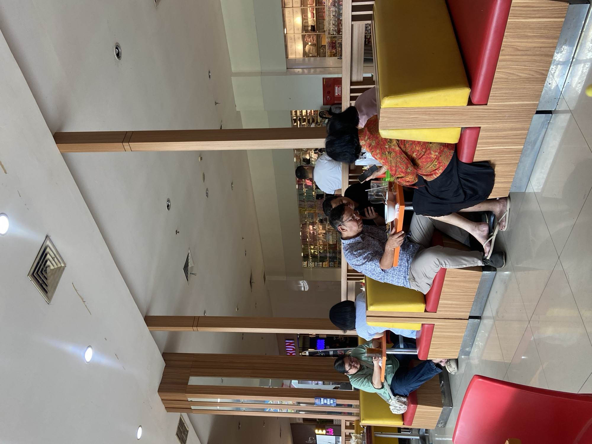 The image shows a group of people sitting at tables in what appears to be a food court or restaurant. The setting is indoors with a tiled floor and a white ceiling with some visible vents and lights. Wooden pillars and partitions divide the space. In the foreground, several people are seated at booth-style tables with red and yellow upholstery. One person in a green shirt is looking down at a table with a drink. Next to them, a person in a blue and white patterned shirt is also looking down, possibly at their phone or food. Further to the right, a woman in a vibrant orange and red patterned top and a black skirt is looking at her phone. Behind them, more people are visible, and in the background, there are displays of merchandise, suggesting a connection to a shopping mall. One sign in the background reads "RUN," possibly part of a store name. The overall atmosphere appears to be casual and relaxed, with people engaged in dining and personal activities. The image quality suggests it was taken with a mobile phone.