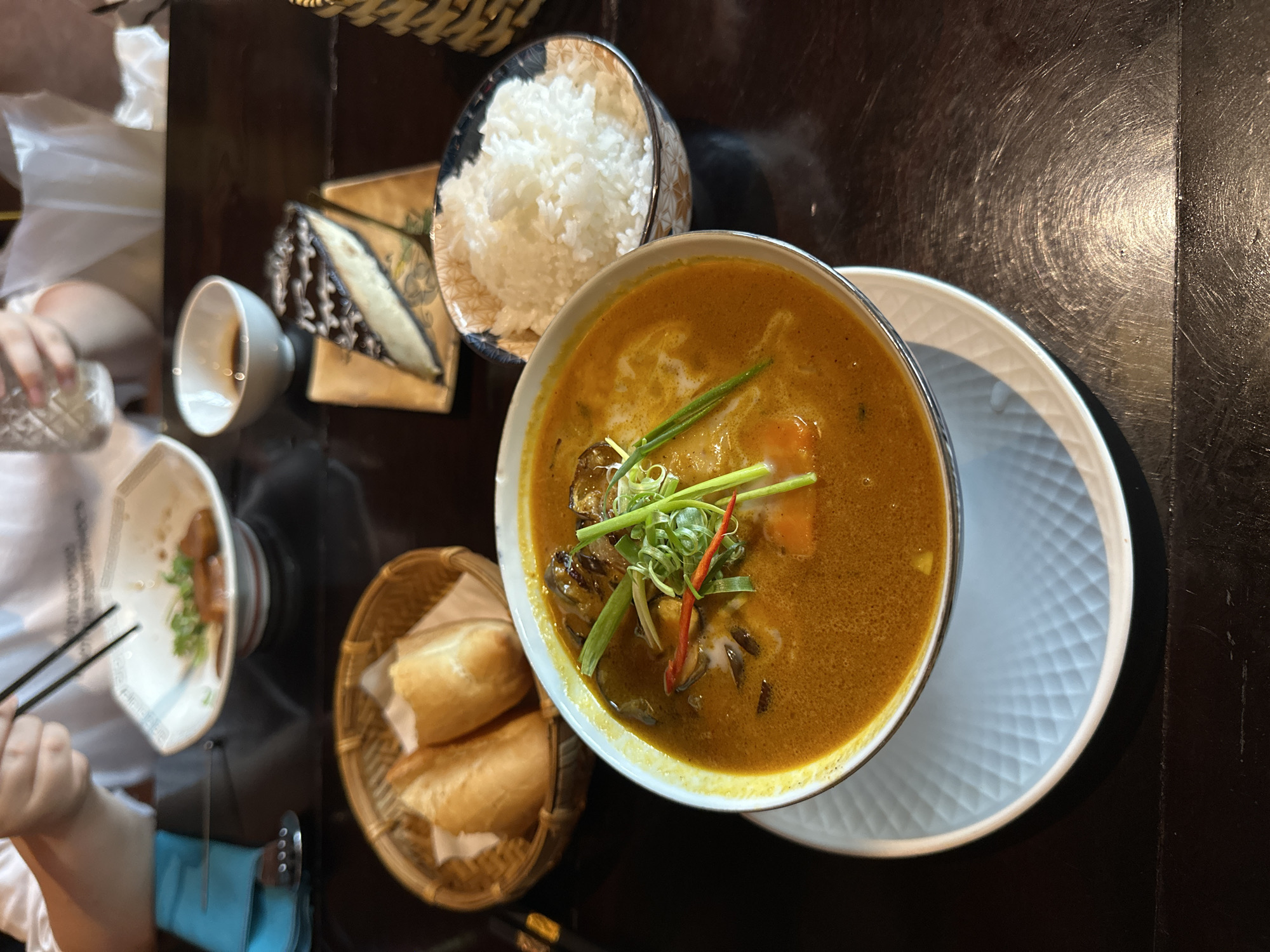 The image captures a close-up view of a table setting in what appears to be a restaurant. The primary focus is a steaming bowl of curry, garnished with green onions and a slice of red chili. To the right of the curry, a small bowl is filled with white rice. In the background, a basket contains two pieces of bread. Further back, a slice of cheesecake with chocolate drizzle sits on a small plate. A white bowl with some food and chopsticks is also visible. A person's hands are partially visible at the top, holding chopsticks and a glass. The table is made of dark wood and the setting suggests a meal is in progress. The overall scene is consistent with dining in a restaurant.
