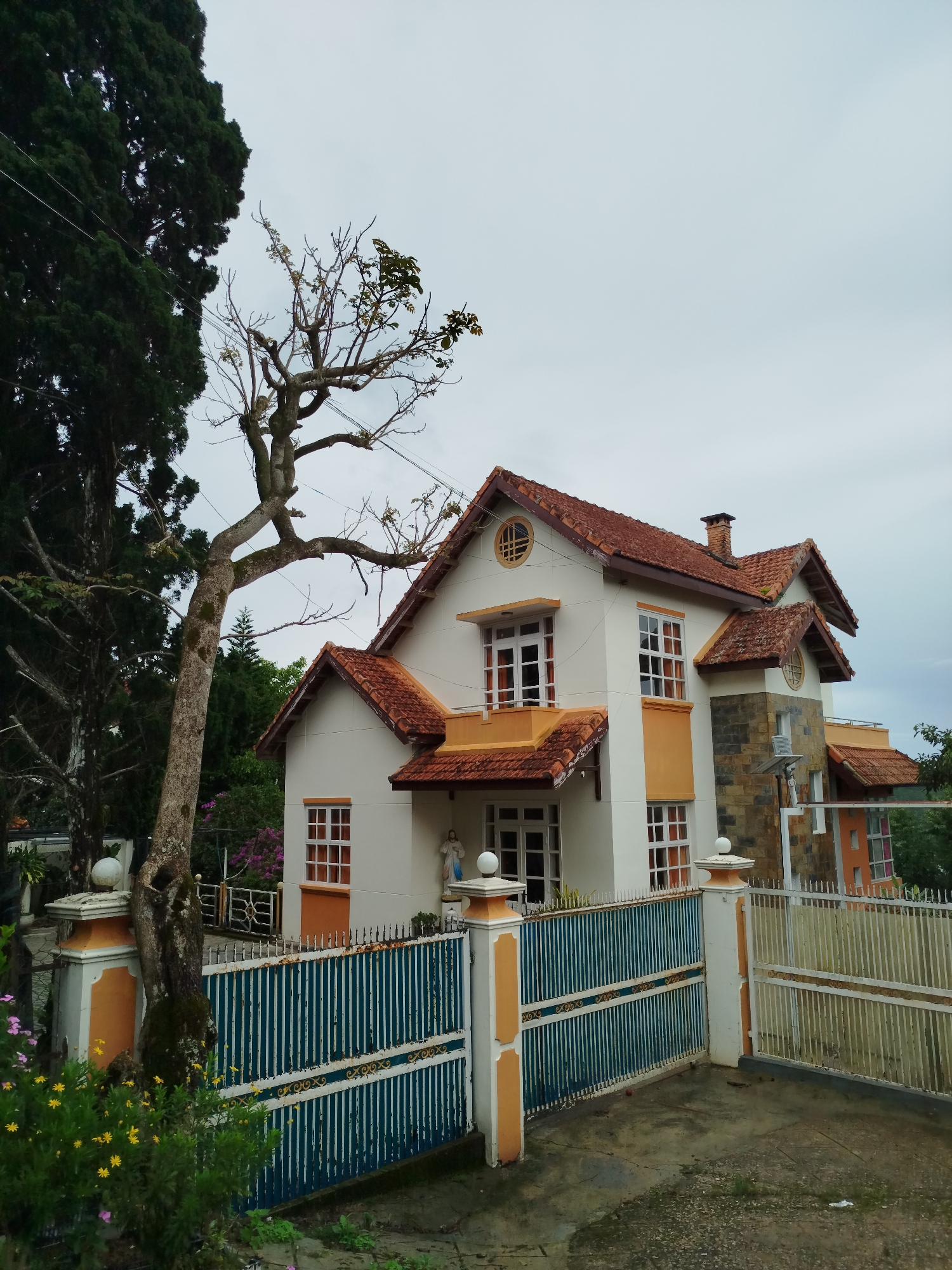 The image depicts a two-story house with a red-tiled roof, white and orange painted exterior, and a stone facade section, located behind a metal fence with blue and white vertical bars and orange posts. A leafless tree stands to the left of the house, partially obscuring the view. A taller, green tree is visible behind it. Yellow flowers grow near the fence. The sky is overcast. The location is in Da Lat, Vietnam.