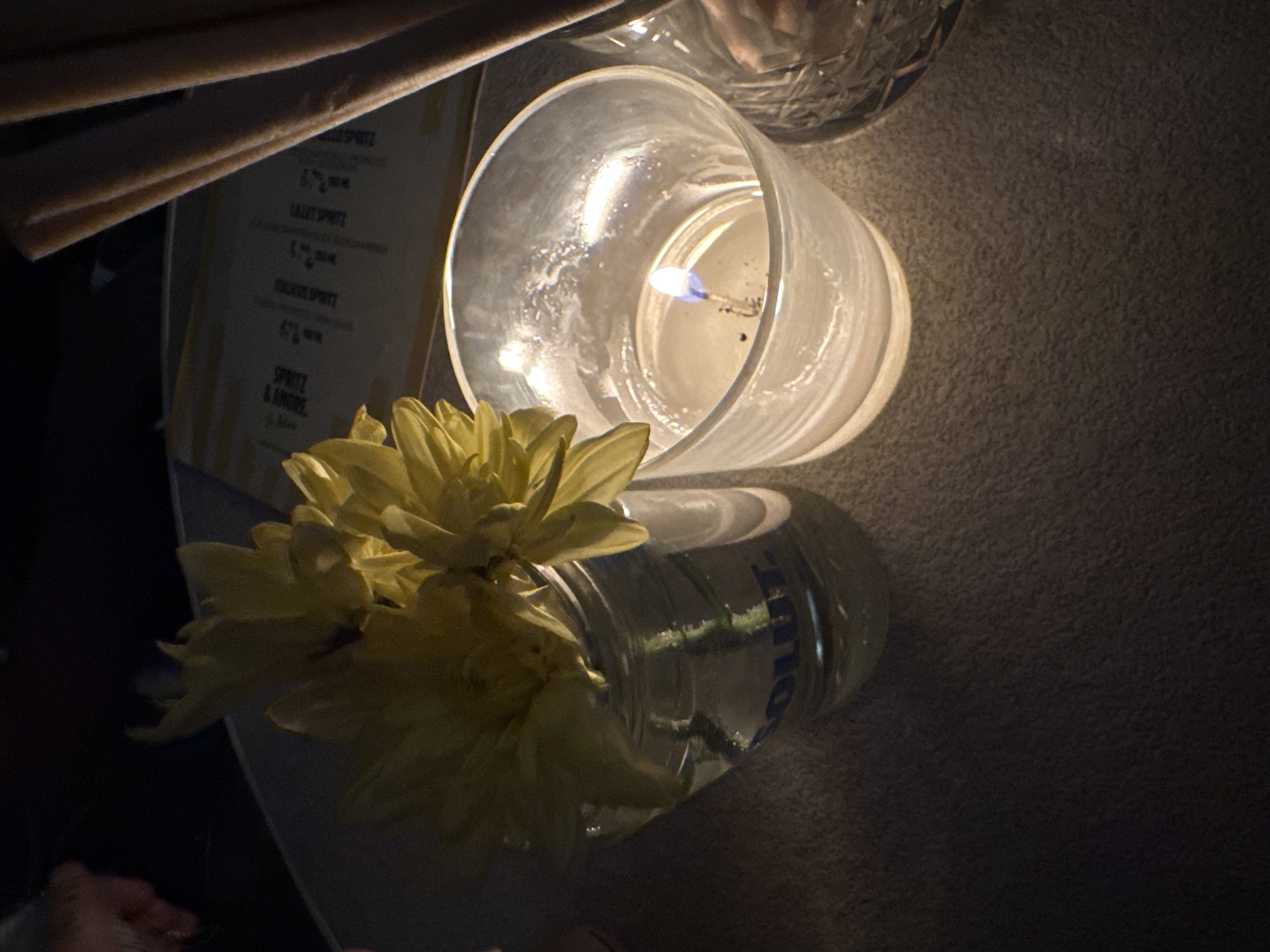 The image shows a close-up view of a table setting with a lit candle in a glass holder, a vase with yellow flowers, and a menu. The candle is burning with a visible flame. The flowers appear to be chrysanthemums or similar, in shades of yellow. The vase seems to be a repurposed Absolut vodka bottle. The background is dimly lit, with a hint of a menu that has text and prices visible, suggesting a restaurant or bar. The overall ambiance is warm and intimate, likely set in the evening. The location is not explicitly stated but is assumed to be Sofia, Bulgaria.