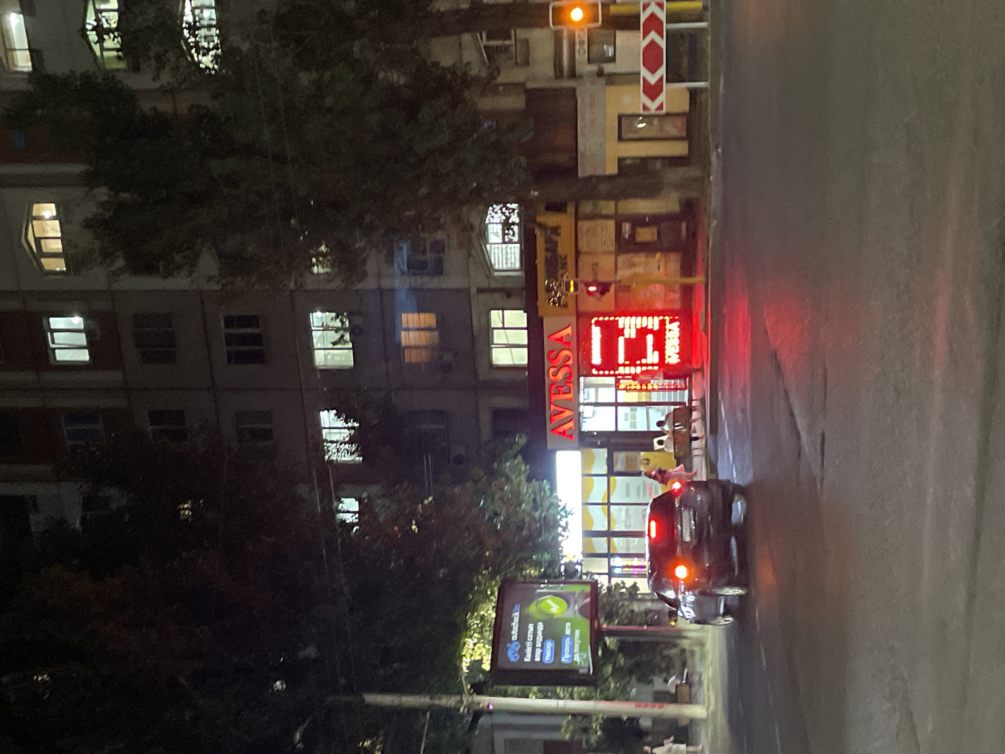 At nighttime on a street in Almaty, Kazakhstan, a dark-colored SUV is driving away from the camera, its brake lights illuminated. To the right of the SUV, a building displays bright red neon signs that spell out "AVESSA" and a series of illuminated vertical lines. Further to the right, a traffic light shows a yellow signal. In the background, a multi-story building with illuminated windows is visible. To the left of the SUV, a billboard displays an advertisement for "autocheck.kz." People are seated on benches outside the building with the neon signs. The street is paved, and trees line the sidewalk.