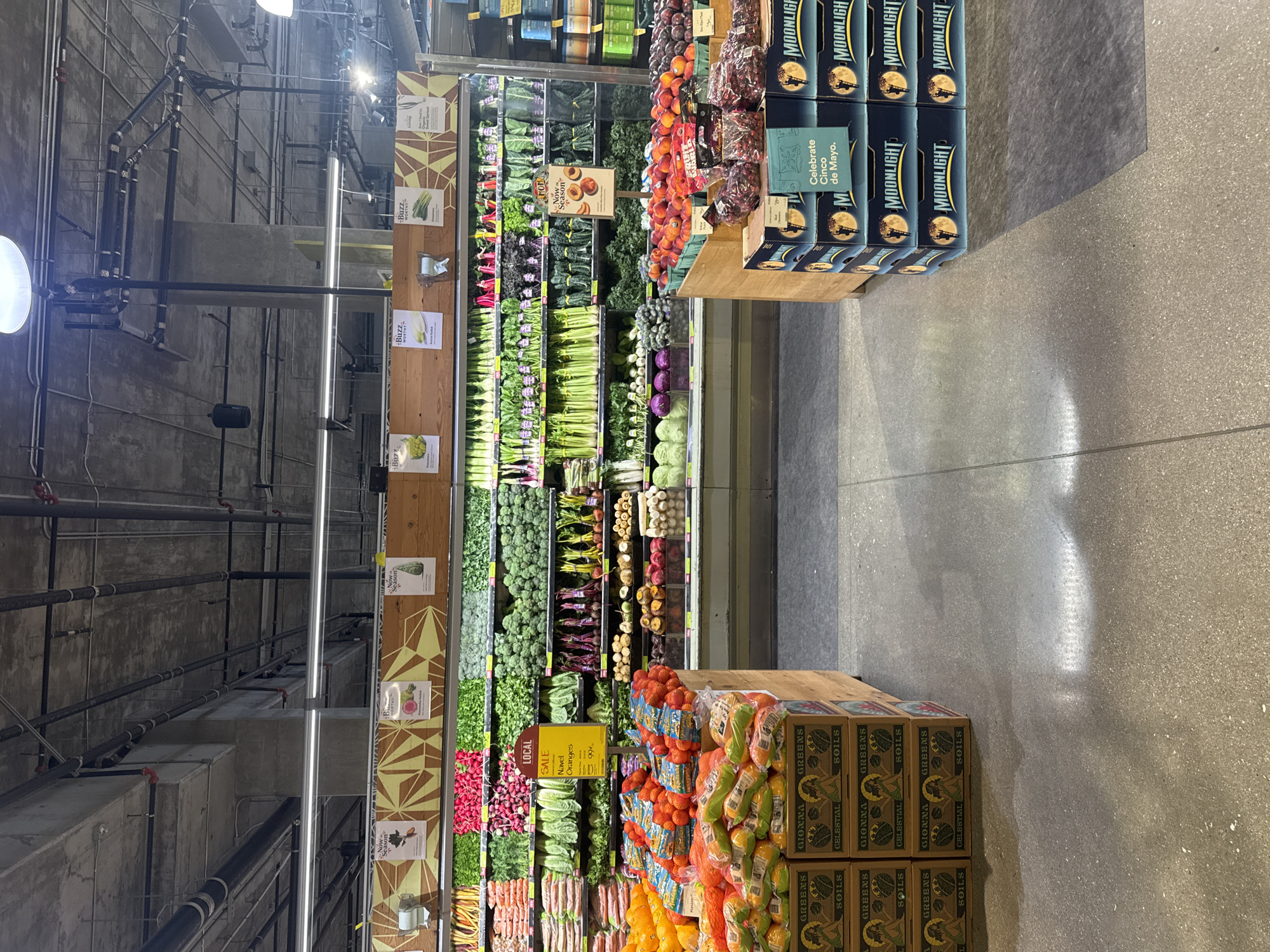 Here is a description of the image:
**Location:** The image appears to be taken inside a grocery store, potentially in Los Angeles, United States.
**Environment:** The area is well-lit, with a focus on the produce section. The background reveals a concrete ceiling structure with exposed pipes and industrial-style lighting.
**Objects & Subjects:**
* The dominant feature is the produce display, stocked with a wide range of fresh vegetables and fruits.
* Boxes of mandarin oranges, some in mesh bags.
* Stacks of boxes of "Moonlight" beverage, labeled with a "Celebrate Cinco de Mayo" design, indicating a possible promotional campaign linked to the Cinco de Mayo holiday.
* Signage showing pricing or labeling for produce, including a "LOCAL SALE Navel Oranges .99" sign.
* "Buzz Worthy" and "Now In Season" labels.
**Actions & Interactions:** The image captures a static scene of a grocery store's display, indicating recent stocking or preparation for shoppers. There is no evidence of human action present.