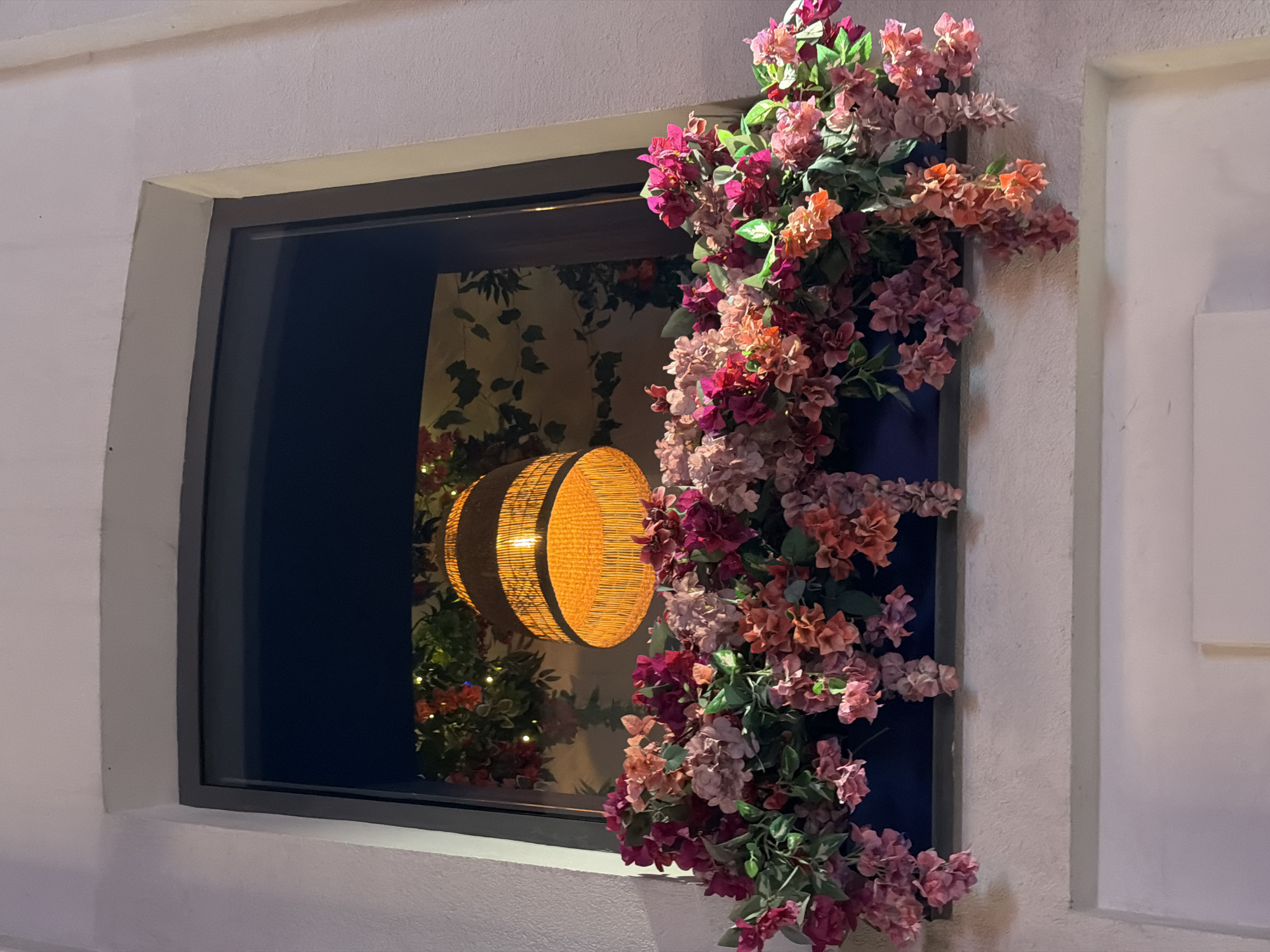 The image shows a window decorated with flowers. The window frame is dark gray and is set into a white wall. A cascade of pink and orange flowers adorns the right side of the window, spilling down from the top corner. Inside the window, a round woven lamp is visible, hanging in front of a wall decorated with foliage. The overall scene has a decorative and inviting feel. There is no indication of this image being in Moscow, Russia.