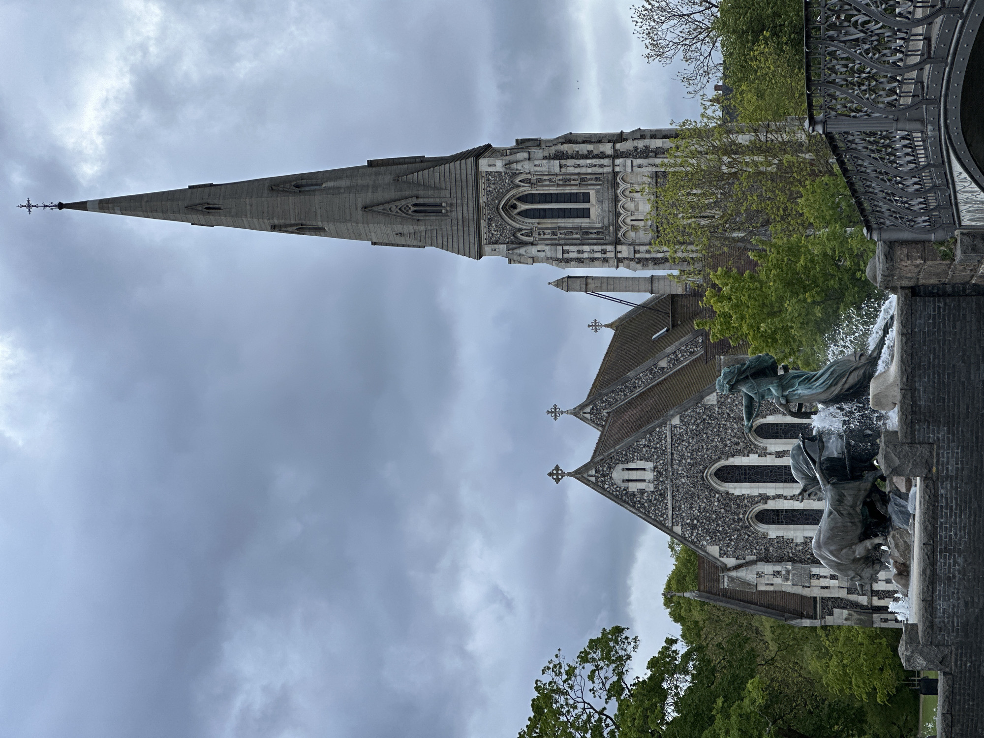 This is a low-angle, outdoor photograph of St. Alban's Church in Copenhagen, Denmark on an overcast day. The towering green steeple of the church is clearly visible. The church's façade includes a Gothic-style window and intricate stonework. To the right of the church is a bridge with ornamental metalwork. In the foreground is a fountain, with figures of animals and water cascading down. Trees with green leaves are visible around the church and fountain, framing the scene. The sky is grey with heavy clouds.