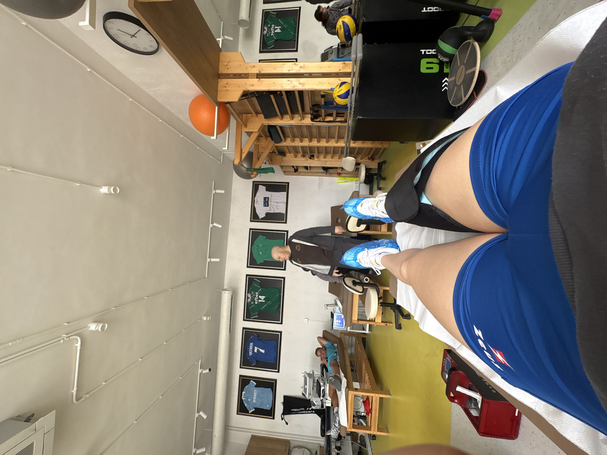 The image presents a first-person perspective from within an indoor physical therapy or sports rehabilitation clinic located in Sofia, Bulgaria. The viewer is positioned on a white-covered treatment table, looking down at their own legs, which are clad in blue athletic shorts and blue and white athletic shoes, with a black support or brace around the upper thigh.
In the foreground, a bald man, likely a therapist or trainer, stands near the patient's feet, observing. Further back, another man in a light blue t-shirt and grey shorts is resting or receiving treatment on a wooden table.
The room features light-colored walls and a yellow-green floor. Numerous pieces of exercise and therapy equipment are scattered throughout, including wooden shelving units holding foam rollers and small exercise balls, two yellow and blue volleyballs, a black apparatus marked "TOC 61", a wooden balance board, and a red machine with "GAMA SPORT" partially visible. A black bag labeled "NORMATEC" rests on the floor.
The walls are adorned with several framed sports jerseys: a green one displaying "ERBET" and "14", a blue one with "VETAROV" and "7", and a light blue jersey featuring "WINBET". A framed white t-shirt and a large circular wall clock showing approximately 12:45 are also visible. Exposed conduit runs along the walls and ceiling, and an orange exercise ball is nestled by a shelving unit. The scene conveys a focused and professional environment for recovery or athletic conditioning.