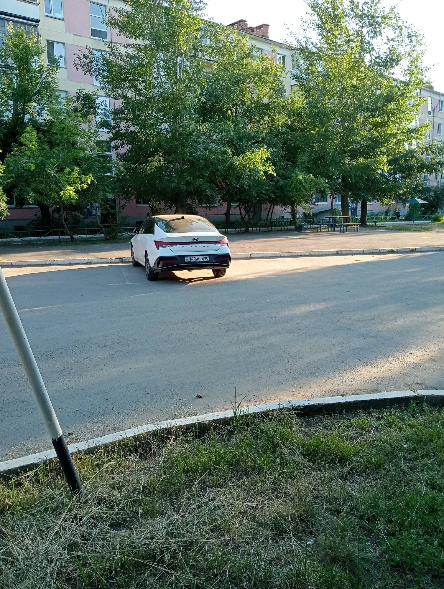 A white Hyundai Elantra is parked on a paved surface in front of a multi-story building. The building has light pink and yellow facades with numerous windows and balconies. Trees with green foliage are situated between the building and the parking area. The ground in the foreground is covered with patchy grass and a concrete curb. A metal pole, partially painted white and black, stands to the left of the frame. The scene appears to be a residential courtyard in a city.
