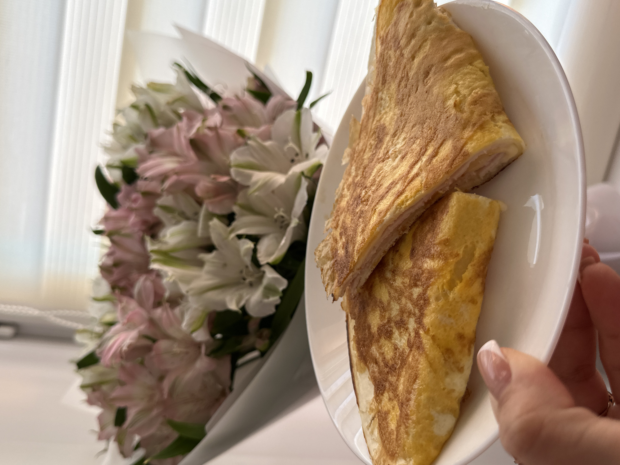 A close-up shot shows a white plate holding two pieces of what appears to be a folded omelet. In the background, a bouquet of pink and white alstroemeria flowers is partially visible, wrapped in white paper. A hand holding the plate is visible in the lower right of the frame. The setting appears to be indoors, possibly near a window with light coming through blinds, based on the vertical lines in the background. Based on the available information, the image appears to be taken in Chaikovsky, Russia.