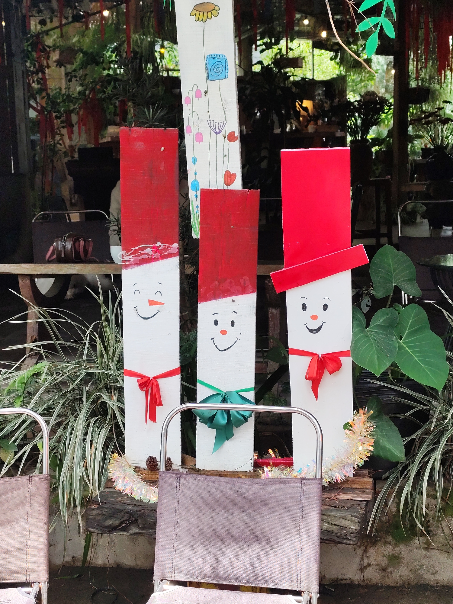 The image depicts a festive Christmas scene in Da Lat, Vietnam. In the foreground, three wooden planks are painted to resemble snowmen. Each snowman has a red painted "hat" on top, a painted face, and a ribbon "bow tie" around the middle. From left to right, the bow ties are red, green, and red. The snowmen are surrounded by plants, and a garland is draped across the bottom. In the background, there is a glimpse of a wooden structure, possibly part of a cafe or outdoor seating area, with hanging plants and other decorations. Two folding chairs are visible in the lower part of the frame, suggesting an outdoor setting. The image exudes a warm, holiday atmosphere with a touch of local charm.