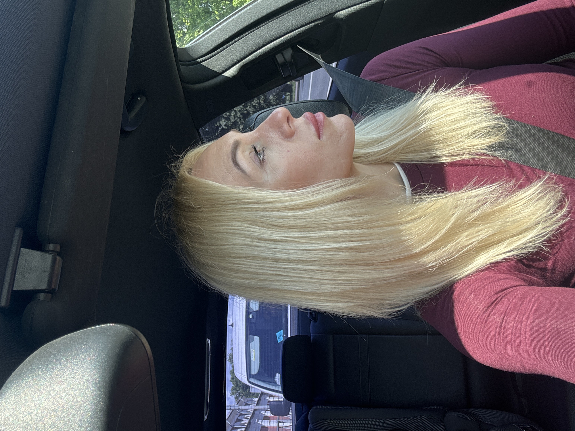 A blonde woman is seated in the driver's seat of a car, looking to her right. She is wearing a burgundy long-sleeved shirt and a black seatbelt. The interior of the car, including the headliner and seats, is visible. Through the car windows, a street scene is visible, with buildings and trees. The sun is shining, casting shadows. The image is likely taken in the city of Saint Petersburg, Russia, given the architecture of the buildings and the style of the vehicles.