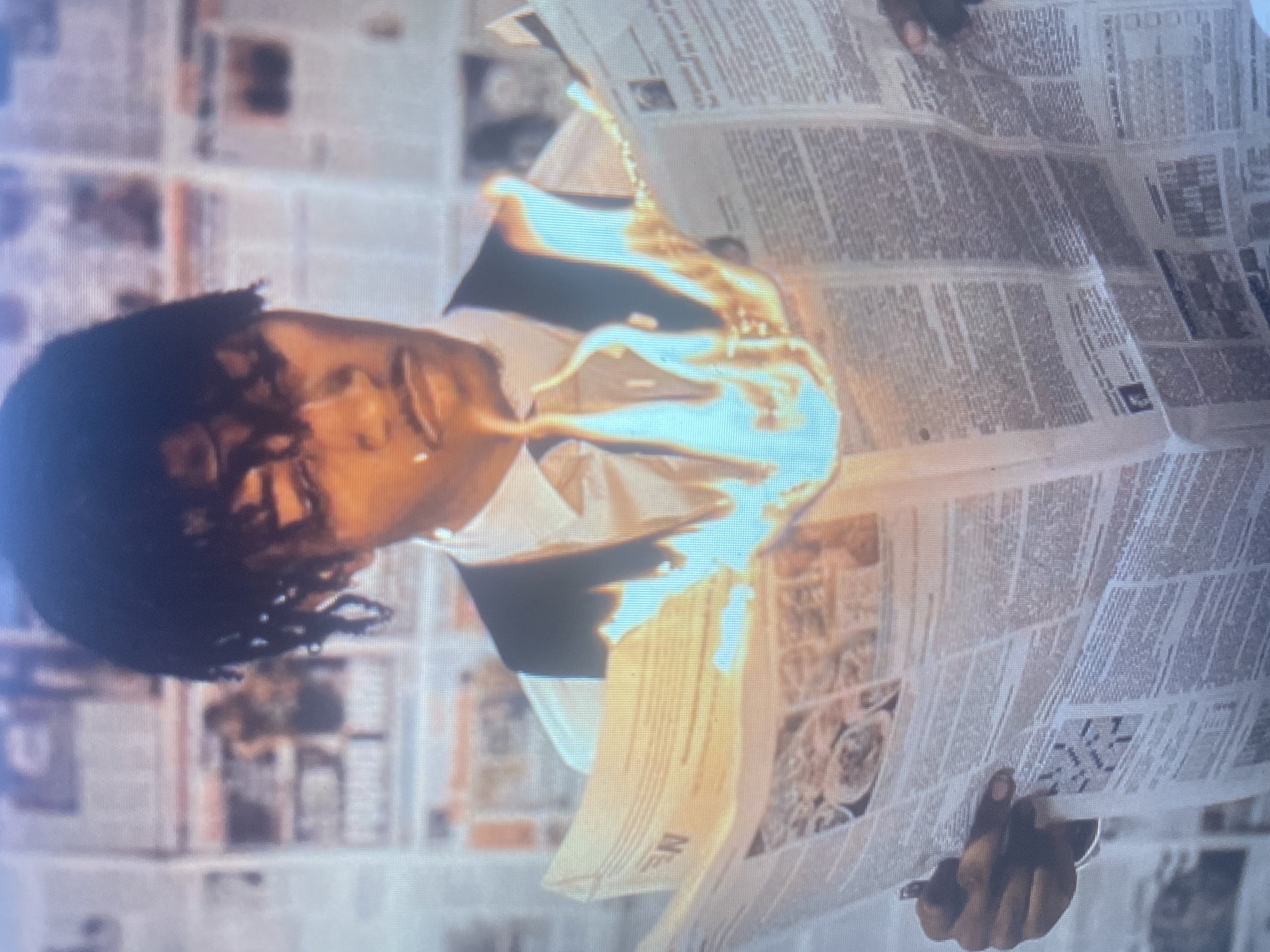 A person with dark, dreadlock-style hair, wearing a white collared shirt and a dark vest, is holding a burning newspaper. The flames are bright orange and blue, and they are concentrated in the center of the newspaper, obscuring some of the text. The person's face is visible, and they are looking down at the newspaper. The background appears to be a wall covered in more newspapers, creating a textured, newsprint-filled environment. The lighting is dramatic, highlighting the flames and the person's face. The person is holding a pen or pencil in their left hand, suggesting they might have been reading or writing before the newspaper caught fire. The scene is set indoors, but the specific location in a city is unknown.