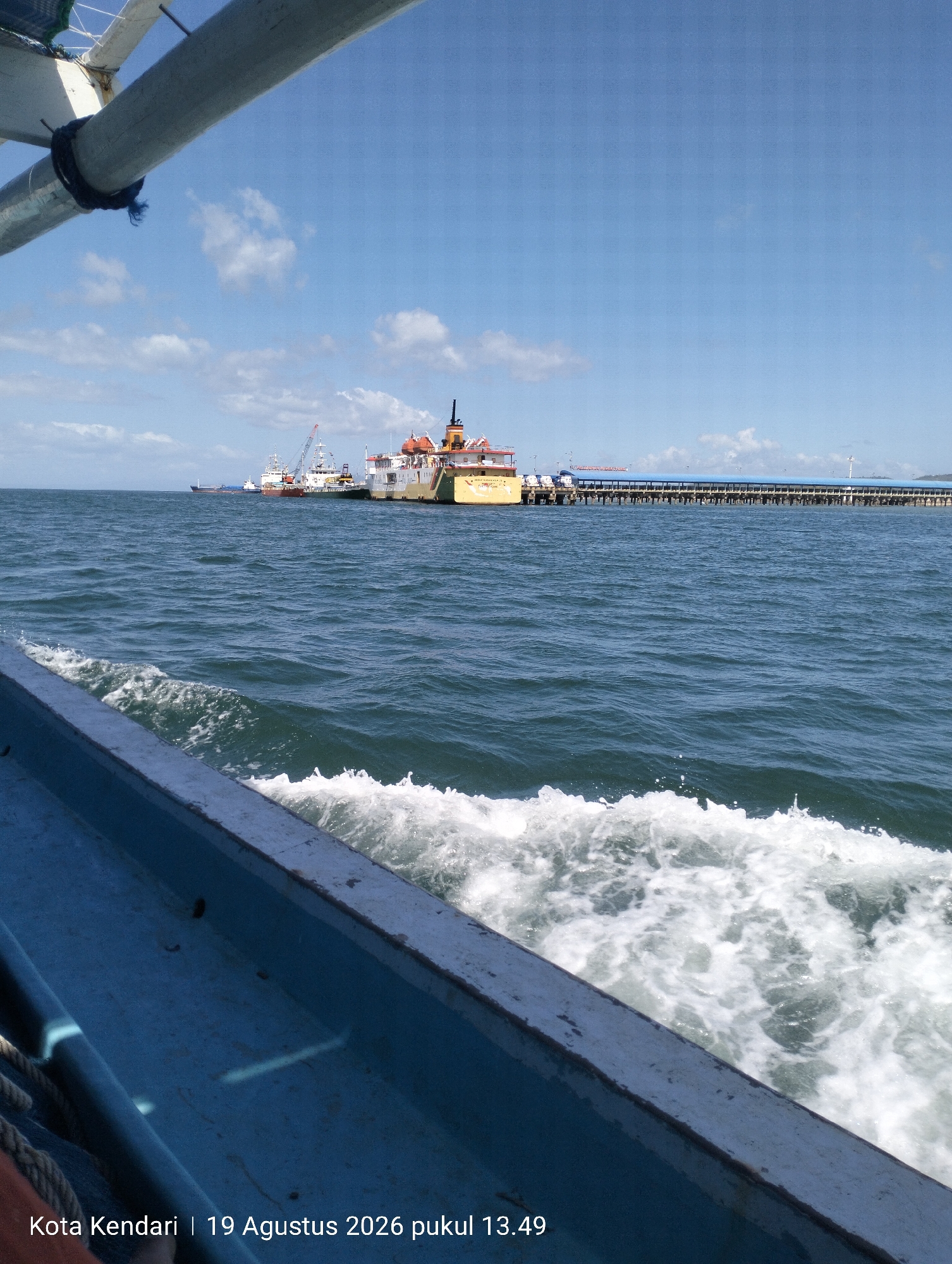 The image shows a view from a boat sailing on the ocean. The boat is in the foreground, with its blue hull visible and waves breaking on its side, creating white foam. In the background, a long pier with a blue roof extends into the water. Several ships, including cargo ships and cranes, are docked near the pier. The sky is blue with scattered white clouds. The text overlay at the bottom of the image reads "Kota Kendari | 19 Agustus 2026 pukul 13.49", which translates to "Kendari City | August 19, 2026 at 13:49". This indicates the location is Kendari, Indonesia, and the image was taken on August 19, 2026, at 1:49 PM.