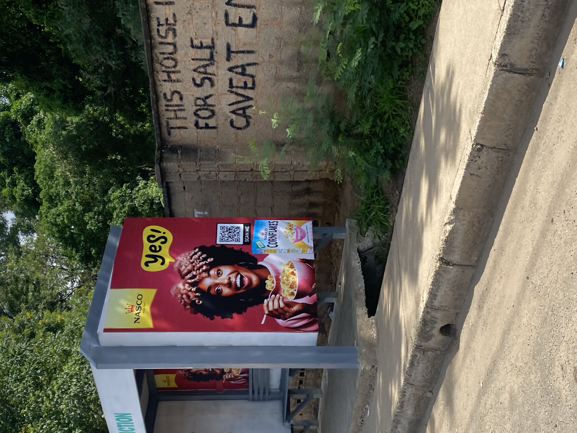The image depicts a street scene in Jos, Nigeria, featuring a bus stop with an advertisement. The advertisement is for Nasco Cornflakes and prominently displays a smiling woman with curly hair holding a bowl of cereal. Next to the woman's image is a box of Nasco Cornflakes and a QR code with the text "SCAN ME." To the right of the bus stop, a brick wall is visible with graffiti that reads "THIS HOUSE IS FOR SALE" and "CAVEAT EN." The bus stop itself is a simple structure with a bench. The surrounding environment includes a paved road, a concrete sidewalk with a drain, and lush green vegetation and trees. The overall scene suggests an urban environment in Jos.