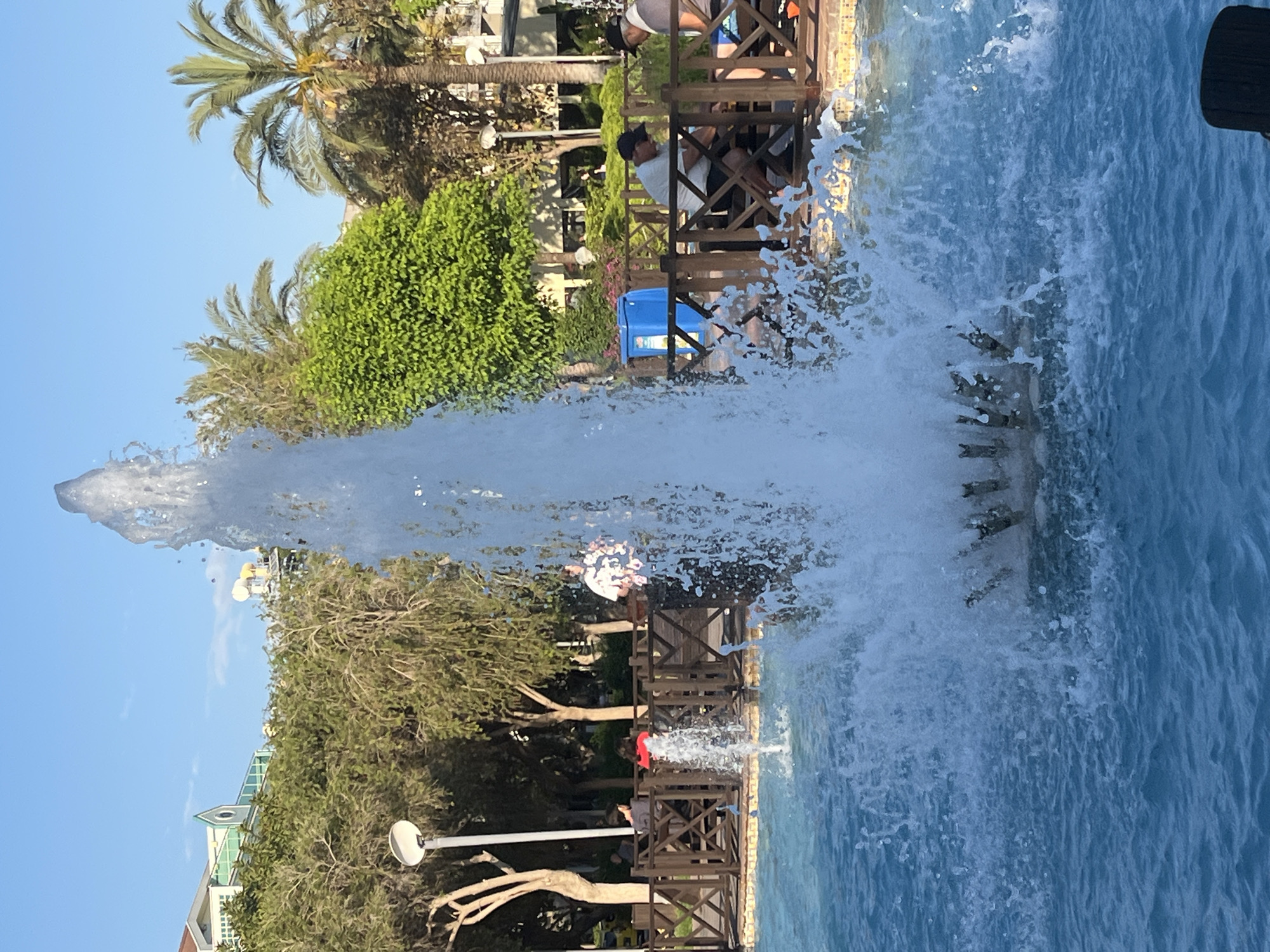 The image depicts a fountain in Alanya, Turkey. The fountain's water jet is the prominent feature, with water spraying high into the air. A blue pool of water surrounds the fountain. Behind the fountain is a wooden railing or fence with people sitting and standing near it. There are trees and palm trees visible, as well as buildings and blue sky. People are observed sitting near the railing and a blue bin is visible in the mid-ground. One person in the image is wearing a red shirt, and another is wearing a white shirt.