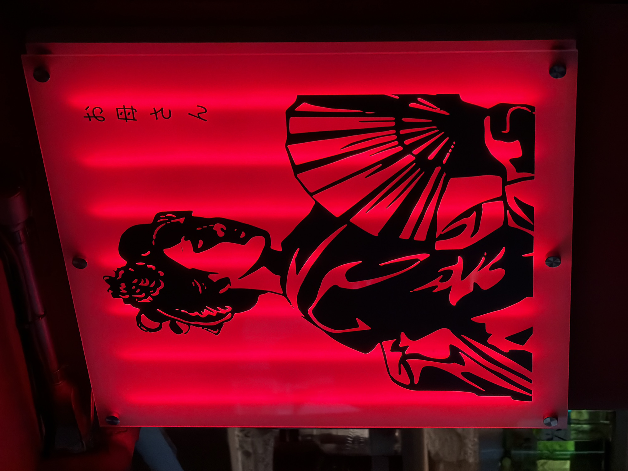 A square, illuminated sign with a black silhouette of a geisha holding a fan against a vibrant red background. The geisha is depicted from the shoulders up, with intricate details in her hair and kimono. To the right of the geisha, in black Japanese script, are the characters "お母さん" (Okāsan), which translates to "Mother." The sign is mounted on a wall and lit from behind, casting a warm, ambient glow. The setting appears to be indoors, possibly a restaurant or bar, with the ambient lighting suggesting a dim environment. The specific location in Bangkok, Thailand cannot be determined from the image alone.