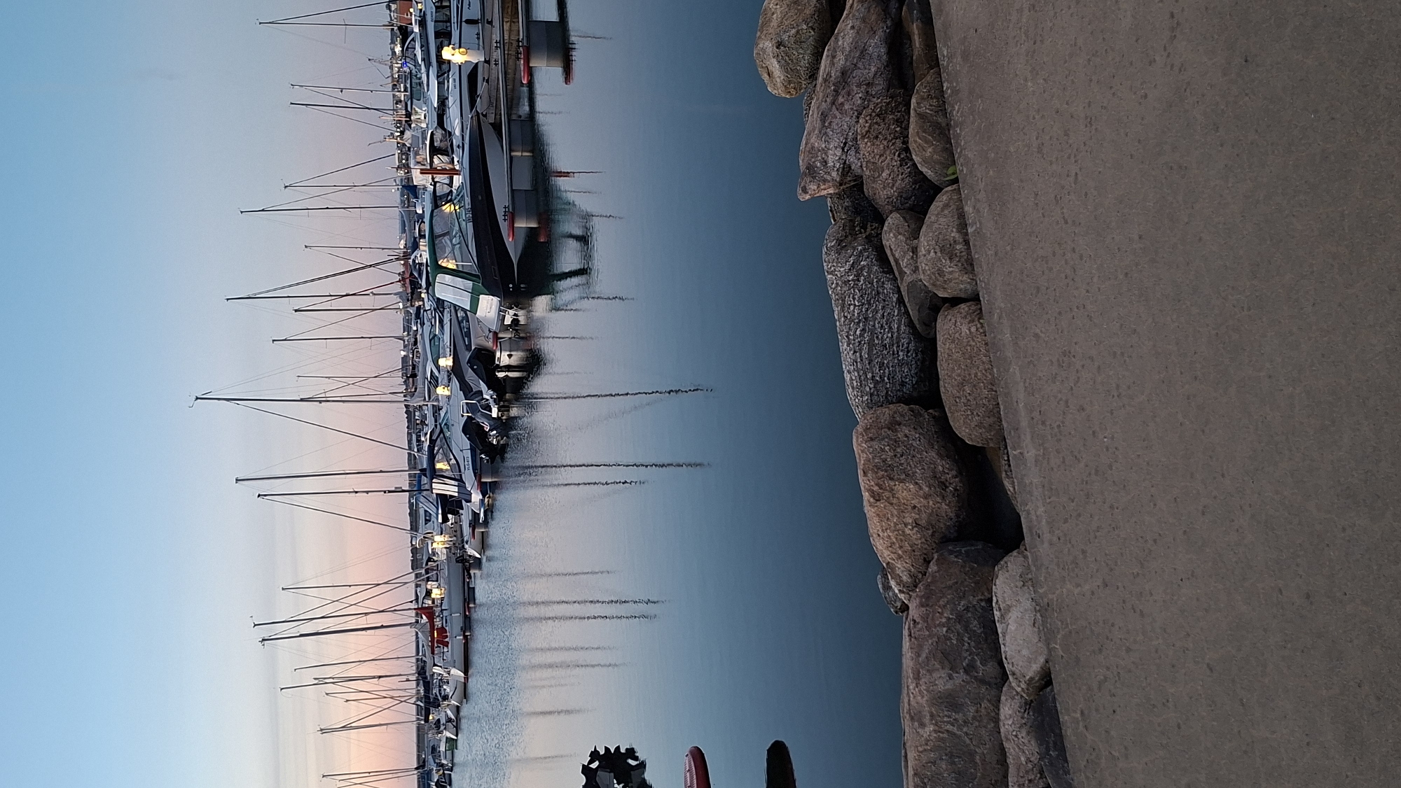This is a vertical, eye-level shot of a harbor in Tallinn, Estonia, at dusk. The sky is a gradient of pale blue to pink, reflecting on the calm water. Numerous sailboats with tall masts are docked in the foreground and middle ground, their reflections shimmering on the surface. Several motorboats are also visible, some with their covers on. The harbor appears well-maintained. Along the immediate foreground, there is a concrete walkway with large, rough-hewn stones lining the edge, separating it from the water. The overall atmosphere is serene and peaceful, suggesting the end of a day at sea.