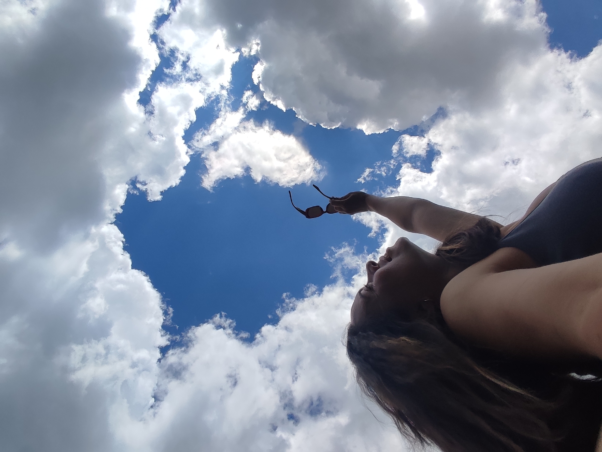 This is an upward-angled outdoor photograph taken in Фоки, Russia, featuring an individual and a partially cloudy sky. The majority of the frame is dominated by a clear blue sky interspersed with numerous white and grey cumulus clouds. A distinct, irregularly shaped patch of deep blue sky, roughly heart-shaped, is prominent in the upper-left quadrant, surrounded by clouds.
In the lower right portion of the image, the profile of an adult individual is visible, oriented to look directly upwards. The person has long, dark, wavy hair that cascades downwards and to the right. Their right arm is extended upwards, and they are holding a pair of dark-framed sunglasses by the bridge against the clear sky. The individual appears to be wearing a dark, sleeveless top or garment. Direct sunlight illuminates the visible side of the person's face and arm, as well as the brighter sections of the clouds, creating shadows within the cloud formations.
