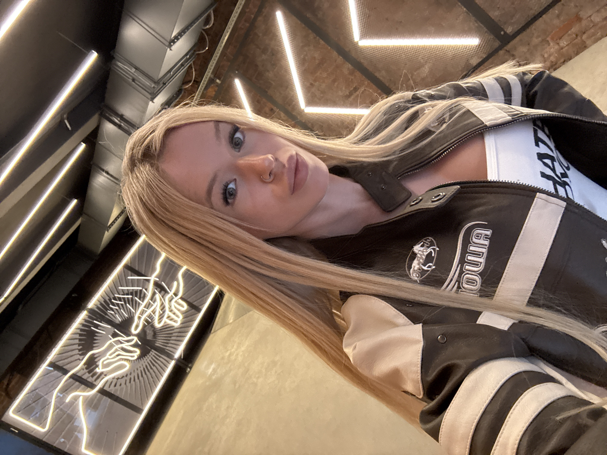 A blonde woman with a nose ring and heavy eye makeup takes a selfie in a dimly lit establishment. She is wearing a black and tan leather jacket with white stripes on the sleeves and a white tank top with black lettering. Behind her, there is a neon sign depicting two hands reaching for each other. The walls appear to be brick, and there are strip lights on the ceiling. The floor is concrete. There is no indication that this location is in Saint Petersburg, Russia.