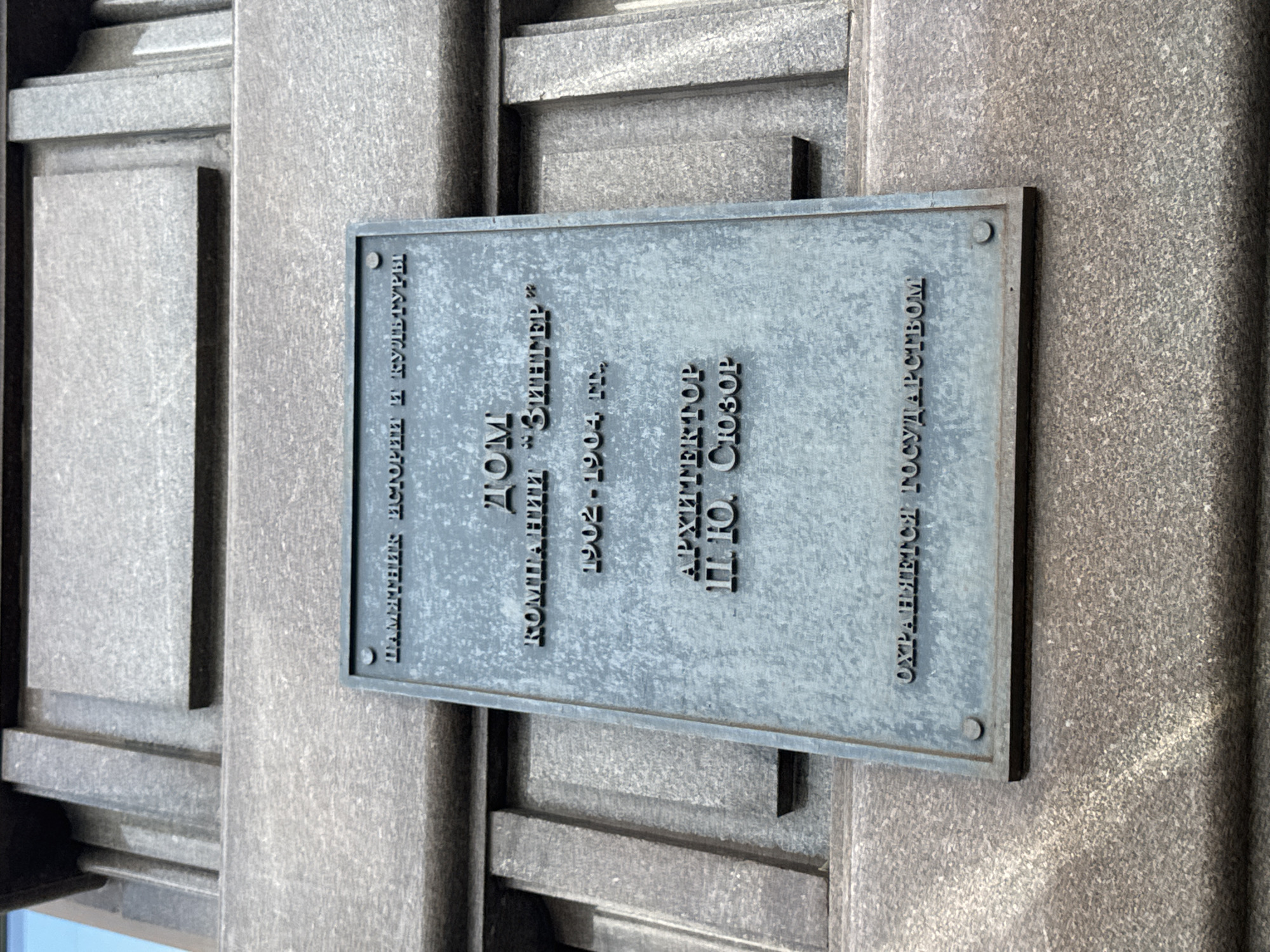 A close-up, low-angle shot shows a bronze plaque mounted on a stone facade in Saint Petersburg, Russia. The plaque is inscribed with text in Cyrillic. The top line reads "ПАМЯТНИК ИСТОРИИ И КУЛЬТУРЫ" (Monument of History and Culture). Below this, it states "ДОМ КОМПАНИИ “ЗИНГЕР”" (Zinger Company House). The years "1902-1904 гг." (1902-1904) are inscribed next. The architect is identified as "АРХИТЕКТОР П. Ю. СЮЗОР" (Architect P. Yu. Syuzor). The bottom line reads "ОХРАНЯЕТСЯ ГОСУДАРСТВОМ" (Protected by the State). The plaque is secured by four visible screws. The stone facade is textured and appears to be part of a building's exterior. The background shows further architectural elements of the building.