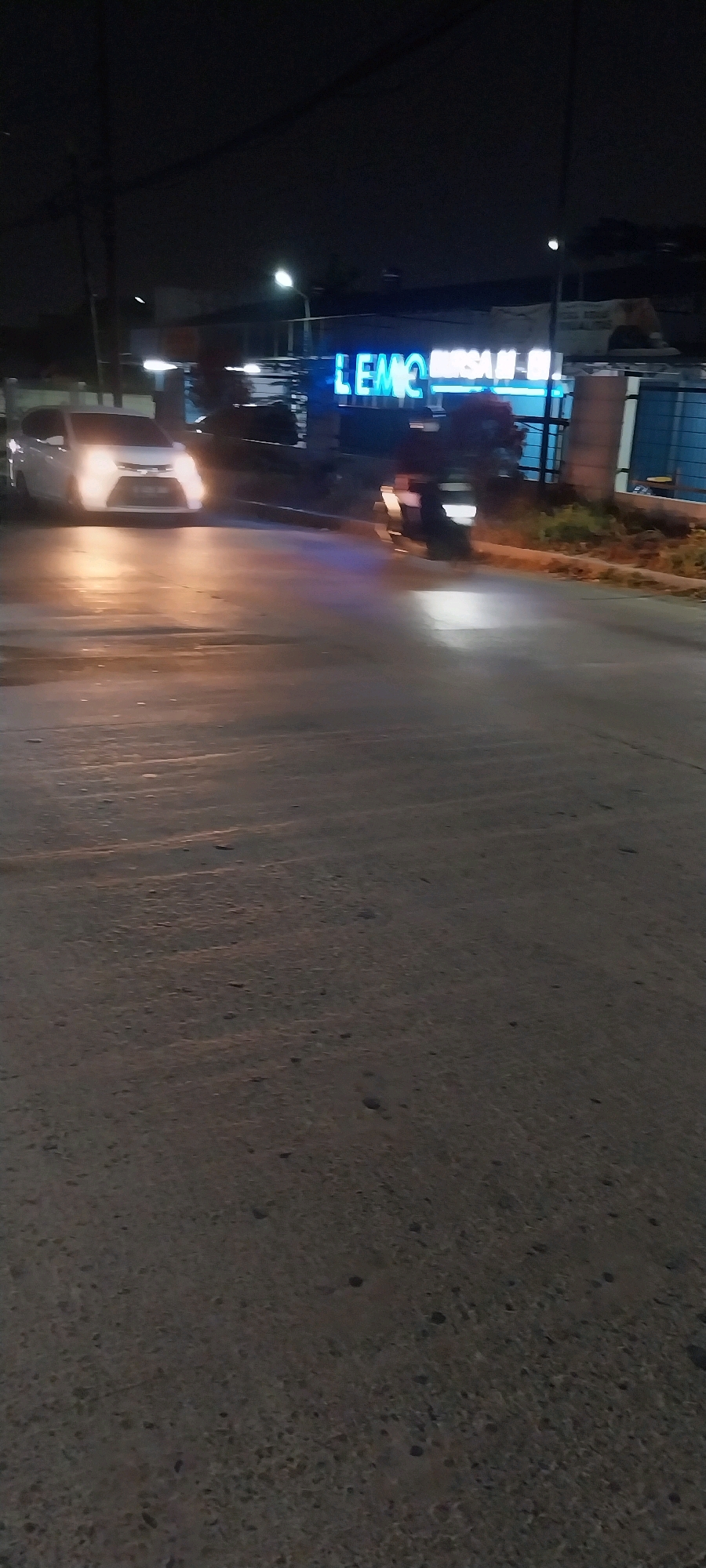At night on a street in Kelapa Dua, Indonesia, a white car is parked on the side of the road with its headlights on, illuminating the wet pavement. A motorcycle is also visible, moving past the car. In the background, a neon blue sign glows, displaying the text "LEMO MRSAI-M". The environment appears to be an urban setting with some vegetation and buildings.