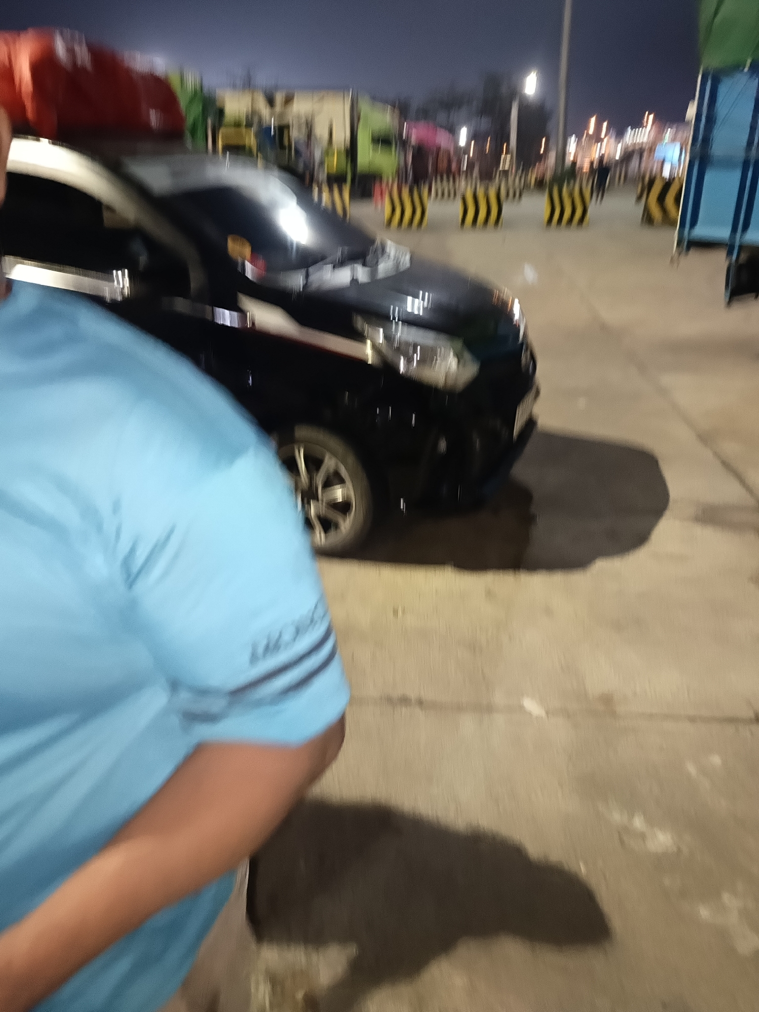 The image depicts a low-light scene, likely at dusk or night, at the Bakauheni Port in Indonesia. In the foreground, a person wearing a light blue t-shirt is partially visible from the shoulder down, facing away from the camera. Behind this person, a black vehicle, possibly an SUV or MPV, is parked on a paved surface. The vehicle's headlights are on, casting reflections on its front. The background features a row of yellow and black striped traffic barriers, suggesting a controlled area. Beyond the barriers, several trucks and buildings are faintly visible, along with streetlights and other illuminated structures, indicating an active port or transportation hub. The overall impression is that of a busy logistical area at night.
