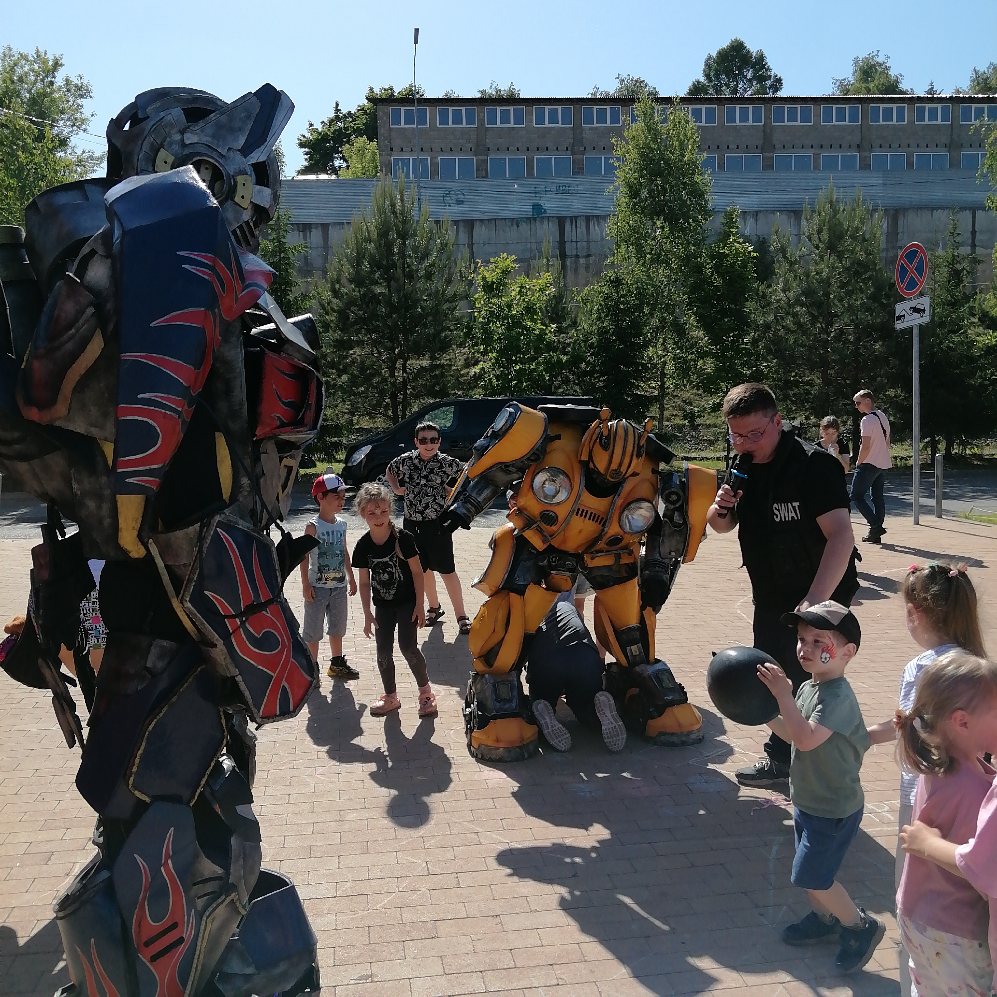 This image captures an outdoor event in Трубачеевка, Russia, under clear skies and bright sunlight, casting distinct shadows on a light-colored brick paved surface.
In the foreground, a person in a large, dark-toned robot costume, resembling Optimus Prime with red and blue flame detailing on black and grey armor, stands on the left, partially cropped, facing right. To its right, centrally located, a person in a yellow and black robot costume, stylized as Bumblebee, is crouched or kneeling, positioned to interact at a lower height.
An adult male, wearing a black "SWAT" vest and clear glasses, stands to the right of the yellow robot, holding a black microphone to his mouth, appearing to address the gathered children. Several children are present: a boy in a white and red cap and blue t-shirt stands near the yellow robot, while a girl in a black t-shirt stands next to him. Behind them, a boy in sunglasses and a patterned shirt observes. In the immediate foreground on the right, a young boy with red face paint (a flame motif) and a dark baseball cap holds a black balloon, looking towards the entertainer. Two other young girls are partially visible to his right.
In the mid-ground, a black van is parked amidst trees. In the background, a large, two-story building constructed of grey concrete blocks with numerous rectangular windows is visible. Below this building, a corrugated metal roof section is present, followed by a concrete wall with faint graffiti. Various green trees are dispersed throughout the background and mid-ground. A tall, dark street light pole stands to the left of the building's upper section. A road sign on the right indicates "No Stopping/Standing" (red circle with blue X) and a supplementary sign showing a tow truck. The scene suggests a public performance or entertainment event for children.