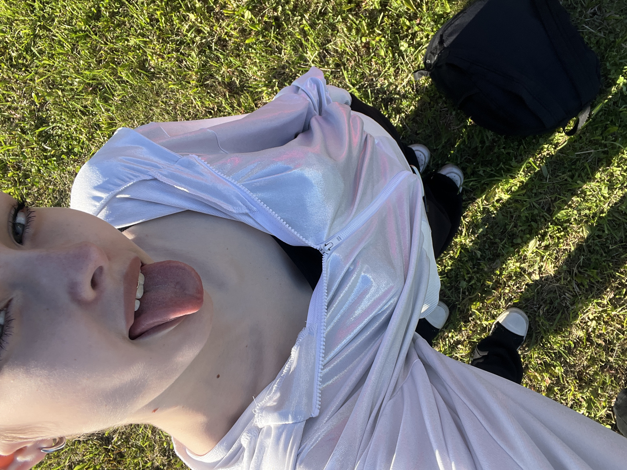 An overhead shot captures a young person from the chest up, lying or leaning back on a grassy surface. Their head is tilted to the left, and they are looking upwards towards the camera with a playful expression, sticking out their tongue. They have light skin and are wearing a light-colored, possibly iridescent or metallic-sheen jacket, partially unzipped, revealing a dark top underneath. A silver hoop earring is visible in their left ear.
The setting is outdoors on a sunny day, evident from the bright natural light and shadows cast on the green grass that covers the ground. In the background, to the right, a black backpack rests on the grass. The person's feet, clad in black shoes with white soles or toes, are also visible on the grass, positioned near the bottom right of the frame. The overall scene suggests a casual and relaxed moment in an outdoor area, possibly a park or field, in Shushary, Russia. There is no visible text in the image.