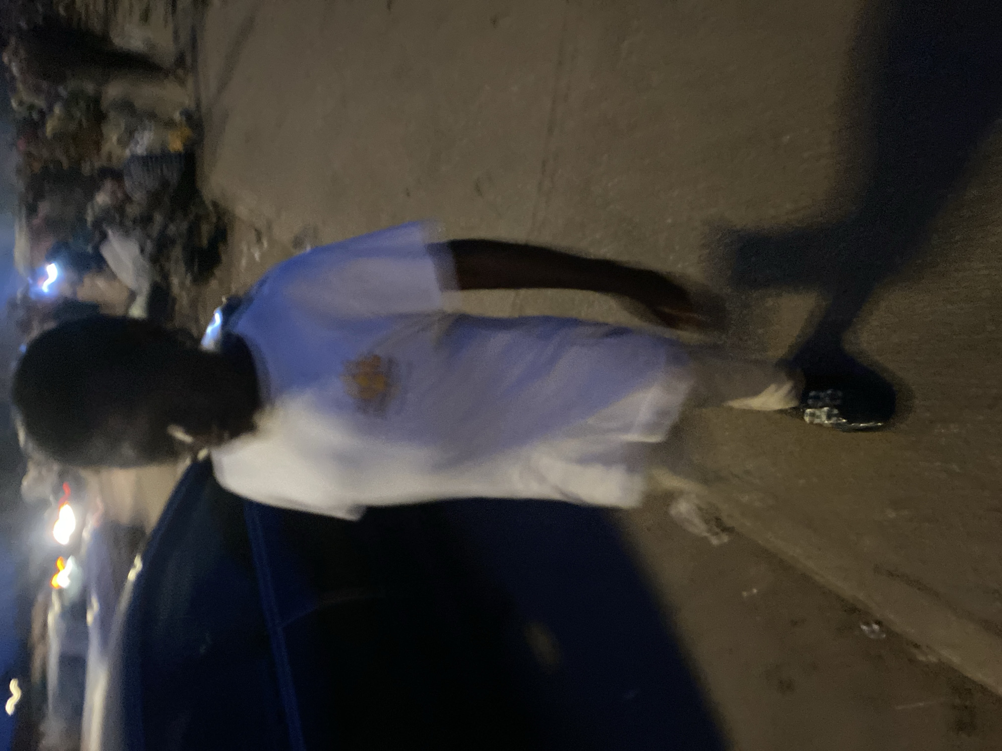 A young individual, appearing to be of African descent, walks on a sidewalk in what is identified as Bukuru, Nigeria. The individual is wearing a white t-shirt with a yellow emblem and dark pants, as well as black shoes. The lighting suggests it is evening or nighttime, with artificial lights from vehicles and possibly streetlights creating a somewhat blurry scene. In the background, there are other people and what appears to be debris or market stalls, indicating a populated urban environment. A dark-colored car is visible on the left side of the image, parked or moving alongside the sidewalk. The ground is paved, with a distinct edge separating the sidewalk from a darker surface, possibly a street or dirt path.