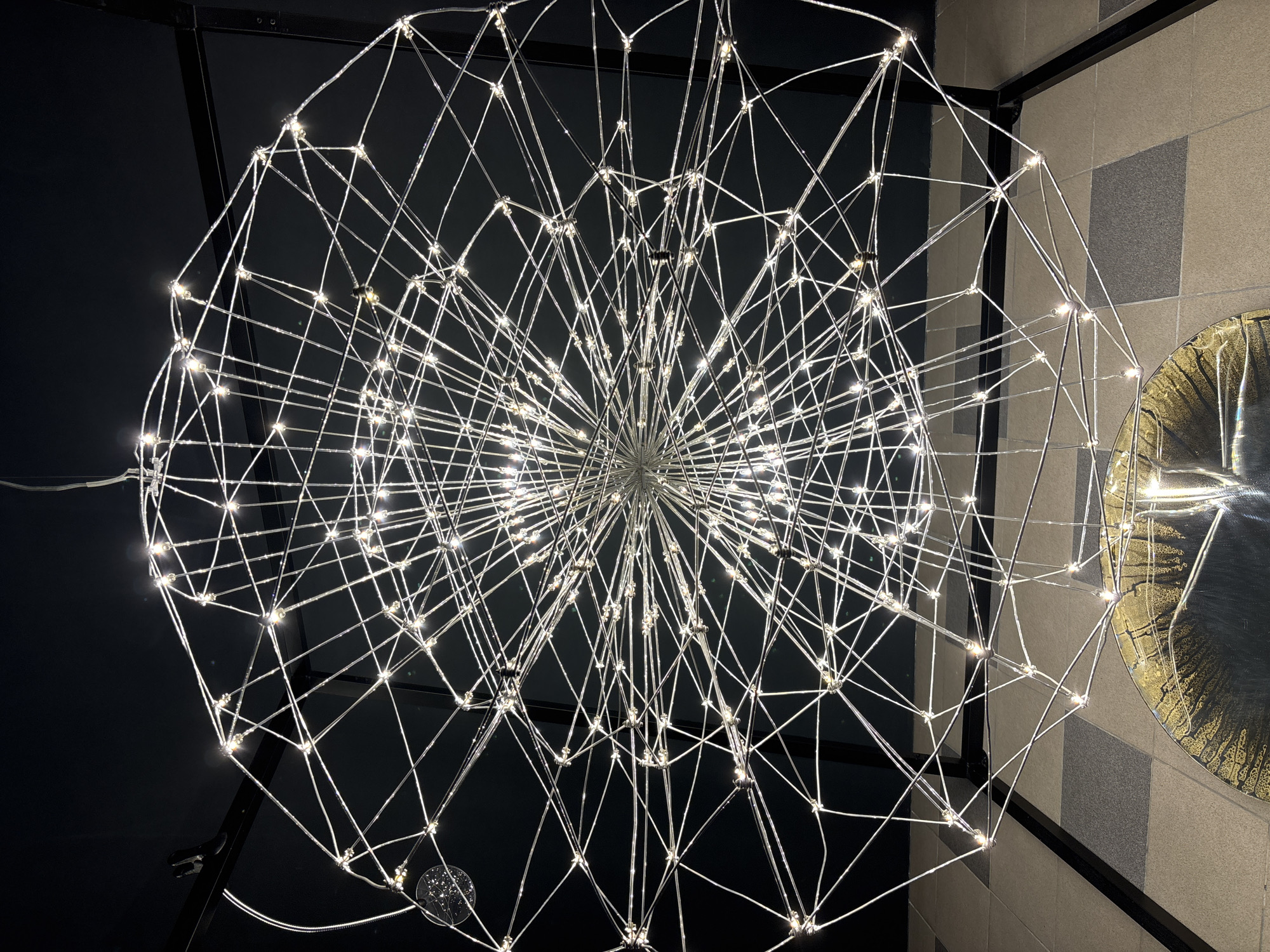 The image shows a decorative light fixture composed of numerous thin, silver-colored metal rods arranged in a spherical structure. At each intersection of the rods, there is a small light emitting a warm, white glow. The background behind the light is split diagonally, with the upper left portion appearing dark, possibly a ceiling or wall in a dimly lit space. The lower right portion of the background is a light-colored wall with a pattern of gray and white squares. A reflective, circular object is partially visible on the right side. The fixture is suspended in what seems to be an indoor environment. This scene is not identifiable as Saint Petersburg, Russia.