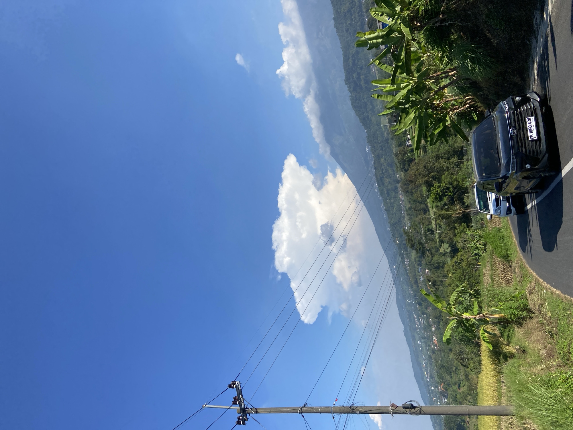 In Sukoreno, Indonesia, a black SUV, identified by the license plate "DK 1850 ACM," navigates a winding road through a lush, mountainous landscape under a bright blue sky. The SUV is in the foreground, closely followed by a white vehicle. To the right of the road, banana trees and other dense vegetation thrive. The road itself is paved and appears to be a two-lane highway. Beyond the immediate roadside, a hillside covered in greenery stretches upwards, dotted with houses that form a village. In the distance, a towering mountain range is visible, with a large, distinct cloud formation resembling a mushroom cloud hovering near its peak. The scene is further characterized by a utility pole with multiple wires extending across the frame, a common sight in rural Indonesian infrastructure. The overall impression is a scenic drive through a vibrant, natural environment.
