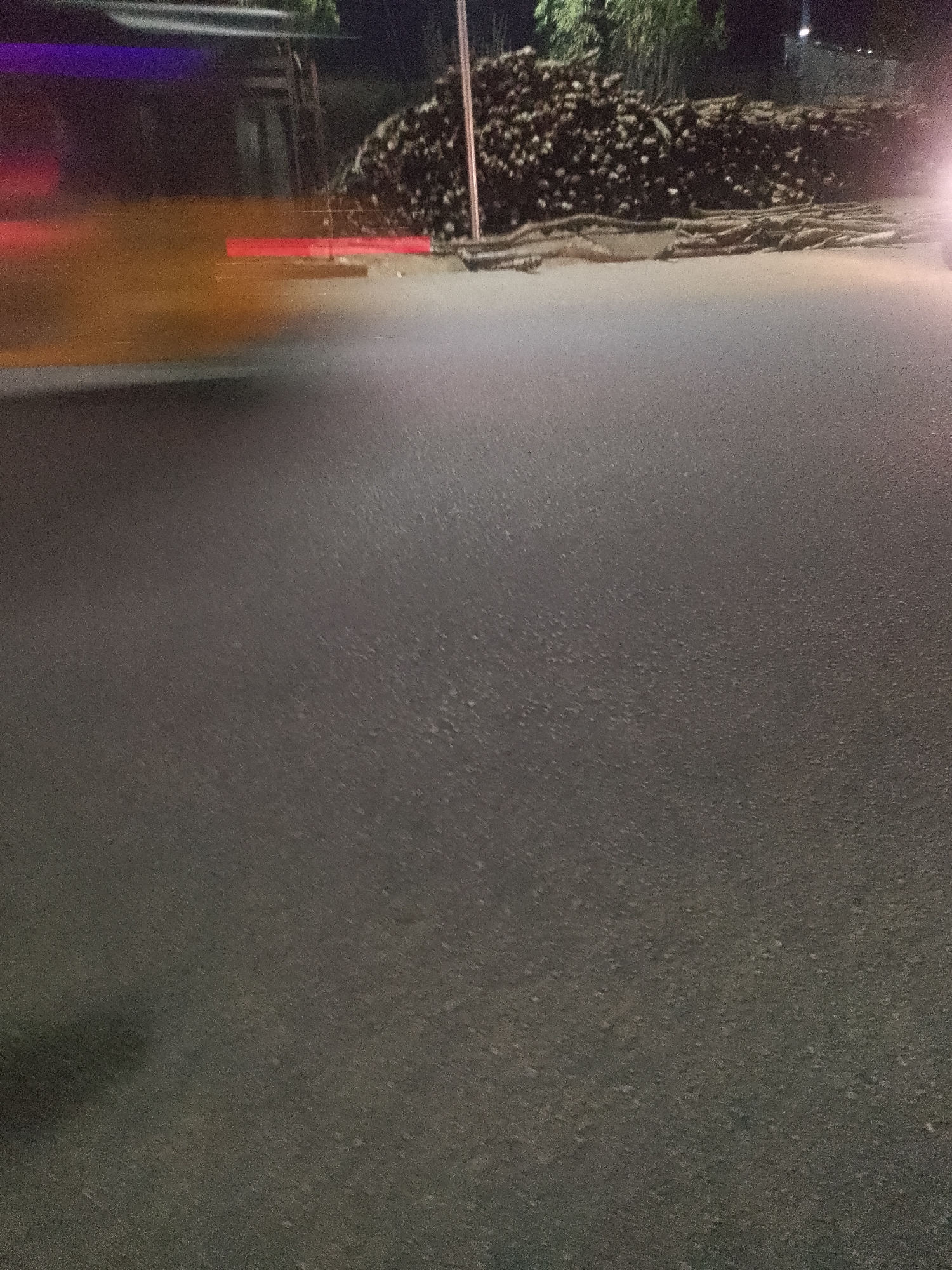 This is a nighttime photograph taken from a low angle, likely from a vehicle in motion, on a paved road in Jalingo, Nigeria. The foreground is dominated by the asphalt surface of the road, which appears somewhat textured. To the left of the frame, there are blurred streaks of light, including a bright purple and a red horizontal band, suggesting movement and possibly streetlights or vehicle lights. In the background, a large pile of what appears to be cut logs or similar debris is visible, illuminated by ambient light. Behind the pile of logs, there are some indistinct structures and foliage, hinting at the urban or semi-urban environment of Jalingo. A bright, out-of-focus light source on the right side of the background suggests another vehicle or a strong light source. The overall impression is of a scene captured during nighttime travel, with motion blur and limited lighting affecting visibility.