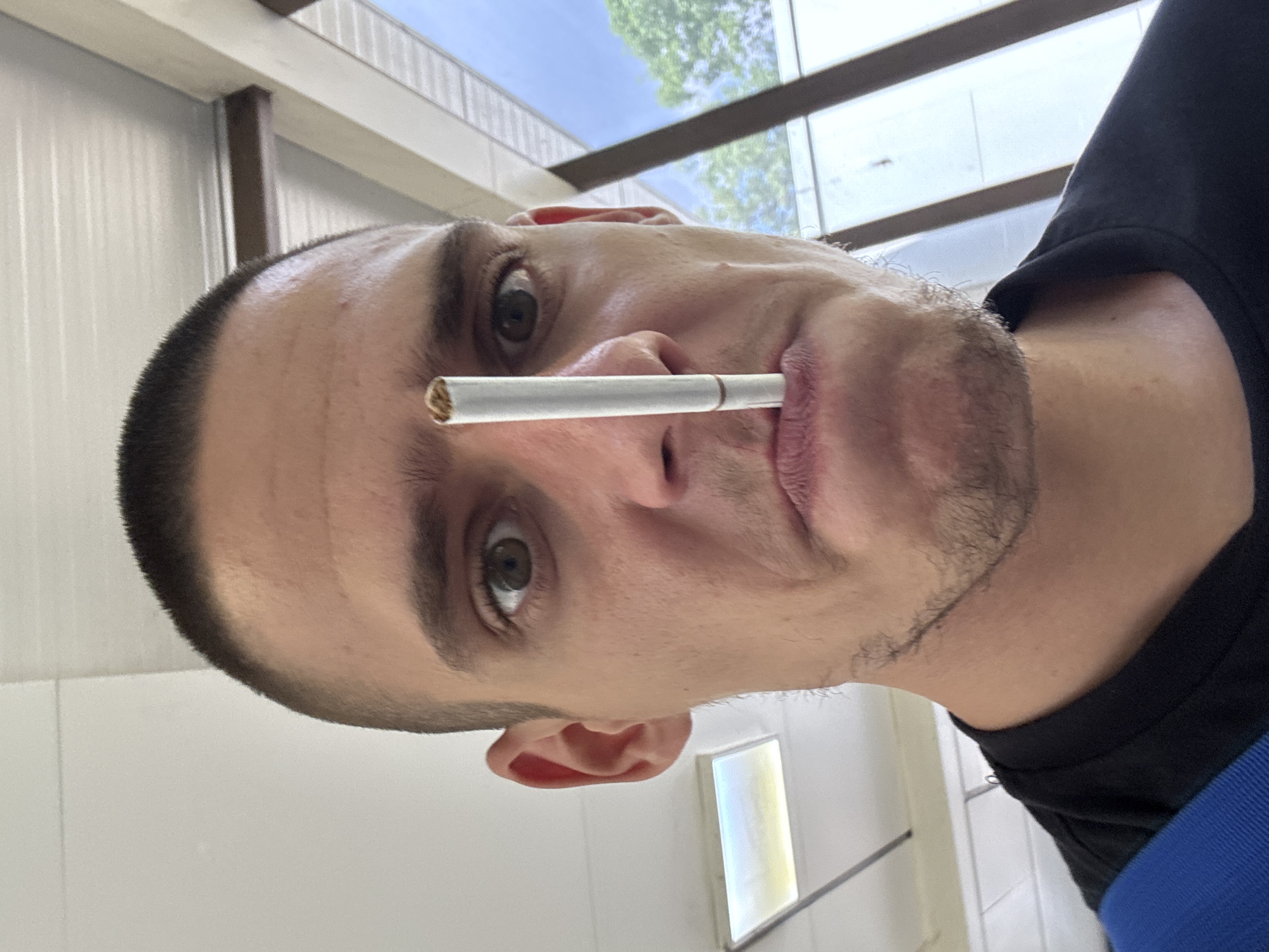 A close-up selfie shows a man with a buzz cut, holding a cigarette vertically in front of his face, with the tip positioned just below his nose and the filter between his lips. He is wearing a black t-shirt with a blue strap visible over his left shoulder. The background appears to be an indoor space with a tiled ceiling and a window with trees visible outside. The image does not provide any information about the location in Sofia, Bulgaria.
