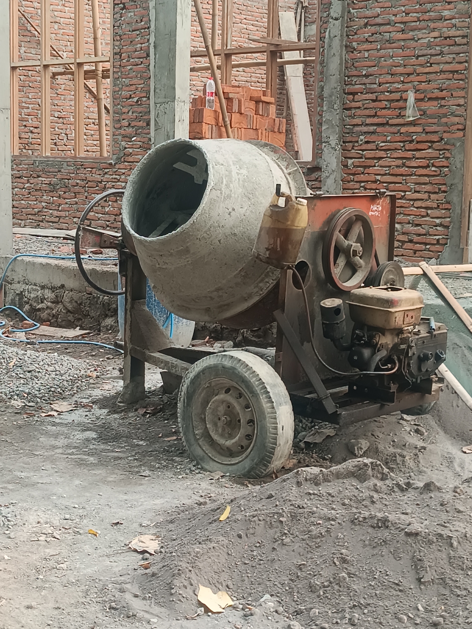 This is a medium shot of a concrete mixer on a construction site in Pleret, Indonesia. The mixer is orange and grey, with a large, worn drum on top. It has a single wheel on the left side and a smaller wheel on the right side. A small engine is attached to the right side of the mixer. The surrounding environment is a dirt ground with piles of sand and gravel, and a partially constructed brick wall in the background. There are also wooden scaffolding and a bottle of water visible. The scene appears to be in the daytime, with overcast lighting. No people are visible in the image.