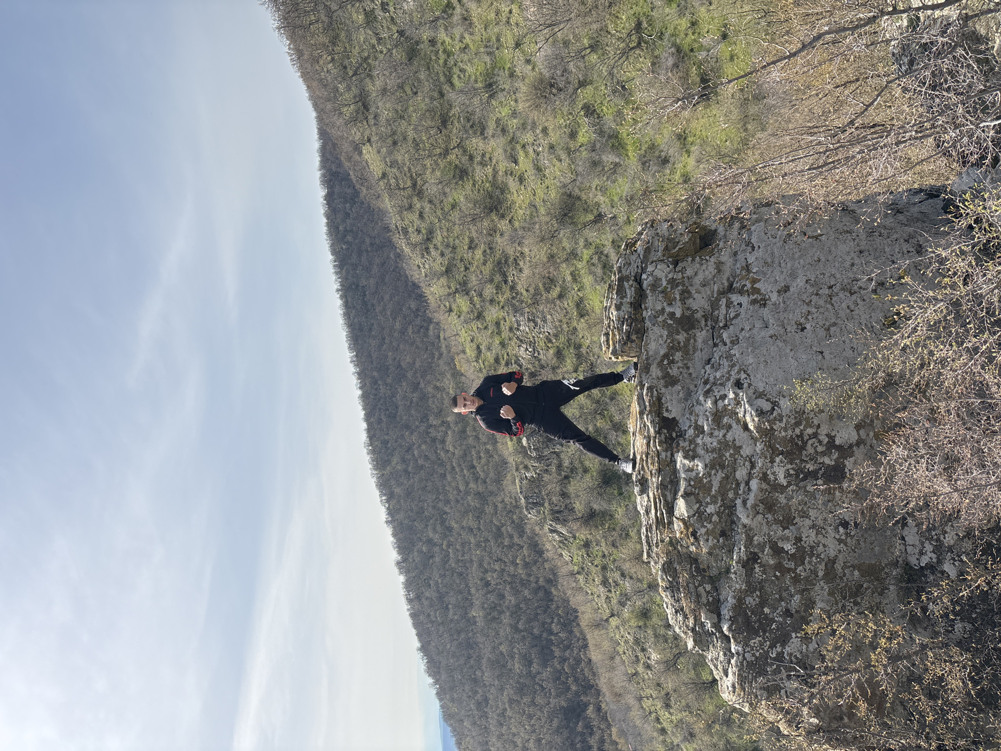 A man is standing on a rocky outcropping, likely a cliff edge, with a forested hillside sloping upwards behind him. The man is dressed in a dark tracksuit with red accents and light-colored shoes. He has his fists raised in a defensive or boxing-like posture. The sky is visible in the distance, appearing light blue and partly cloudy. The photo appears to have been taken outdoors, with natural lighting. The image suggests the location is Goritsa, Bulgaria, based on the prompt.