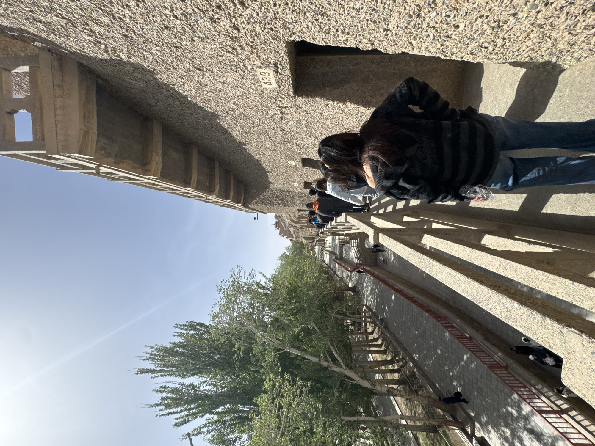 The image depicts a walkway beside the Mogao Grottoes, a UNESCO World Heritage Site near Dunhuang City, China. Several people are standing on the walkway, viewing the grottoes. The walkway is lined with stone structures, trees, and a low red fence. The sky is clear and blue. The stone wall adjacent to the path is labelled "439."