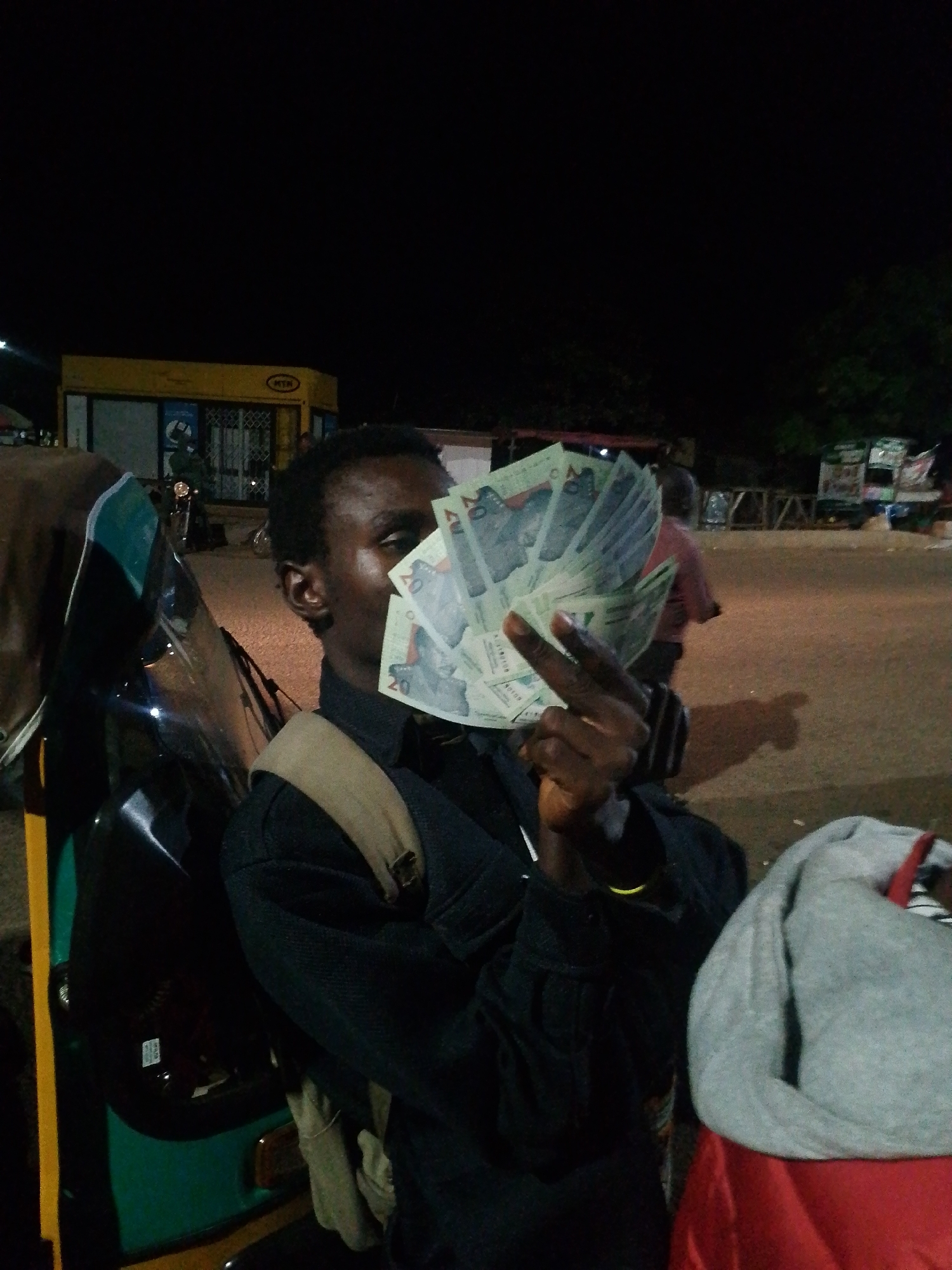A young man, partially obscuring his face with a fan of banknotes, sits in the driver's seat of a three-wheeled vehicle at night. He wears a dark collared shirt with a tan strap across his chest. The vehicle's green and black exterior is visible. In the background, a yellow building with the "MTN" logo is lit, and other vehicles and people are present in the dimly lit outdoor environment.