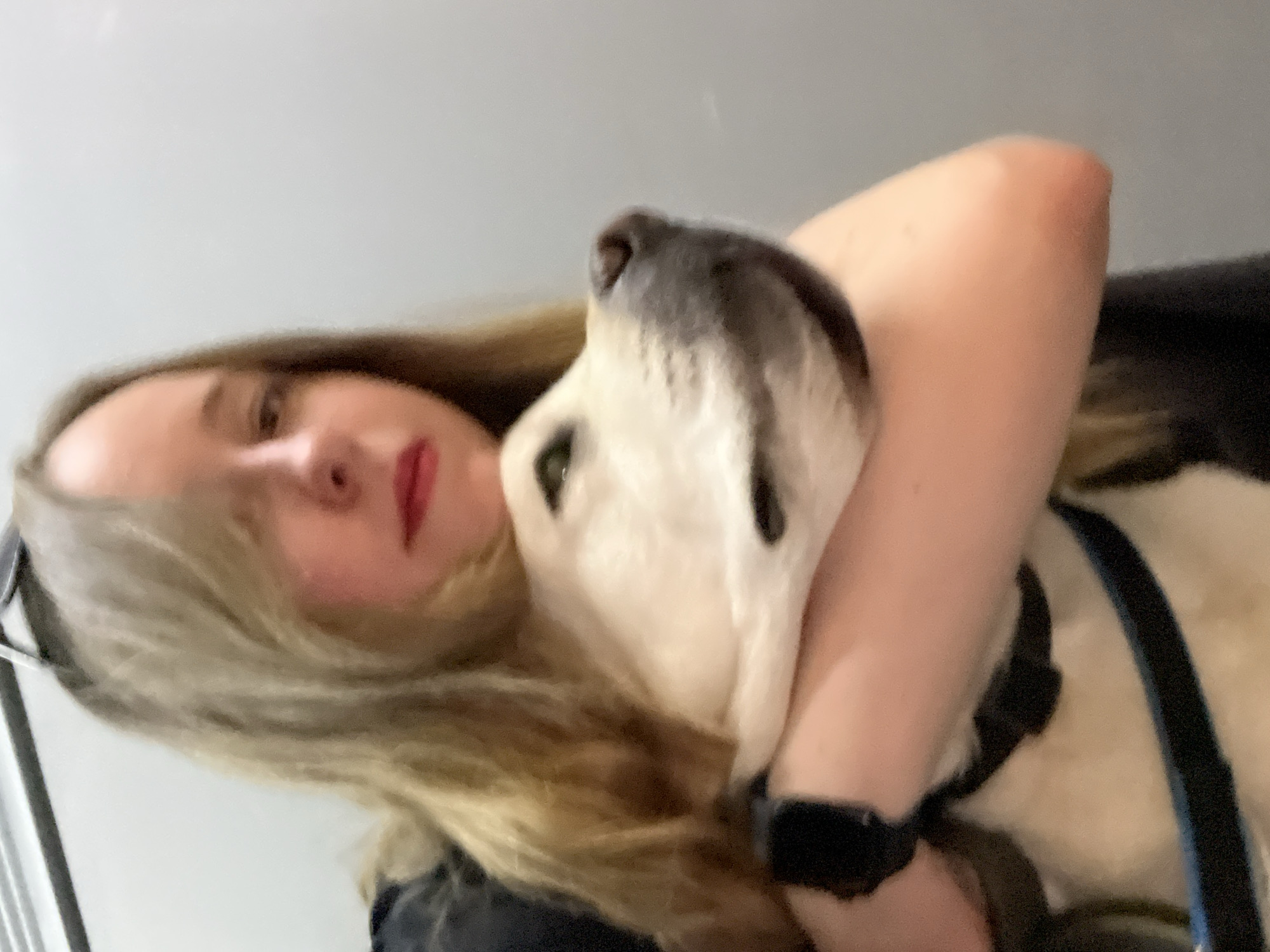 A blurry, rotated, close-up photograph shows a woman with long blonde hair and red lipstick embracing a dog. The dog has white fur and a black muzzle. The woman's arm is wrapped around the dog's neck. A black collar and blue leash are visible on the dog. The background is a plain light gray. No identifiable location in Mytishchi, Russia, is discernible from the image.